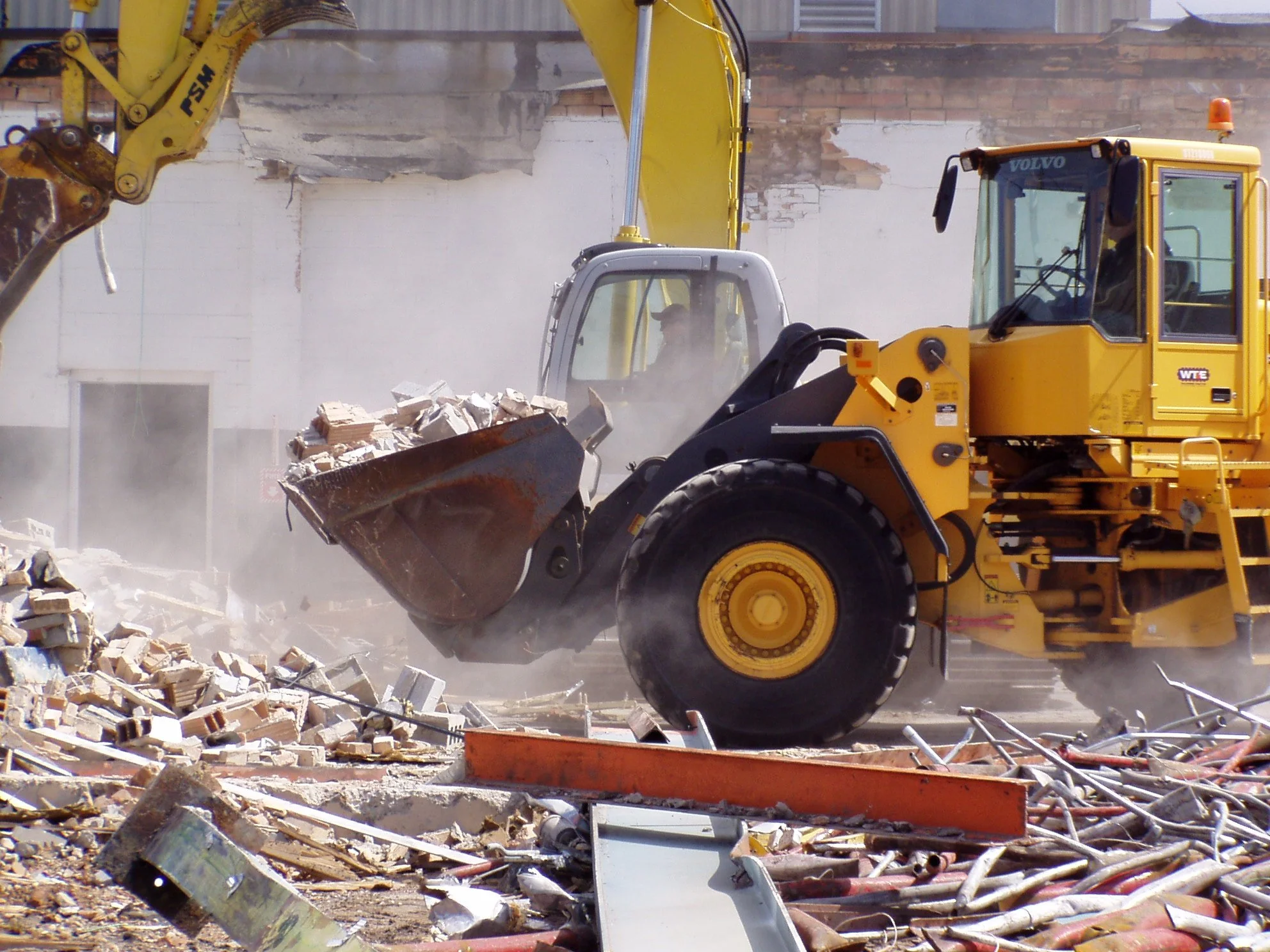 A yellow bulldozer clearing debris and rubble at a demolition site with dust flying in the air.