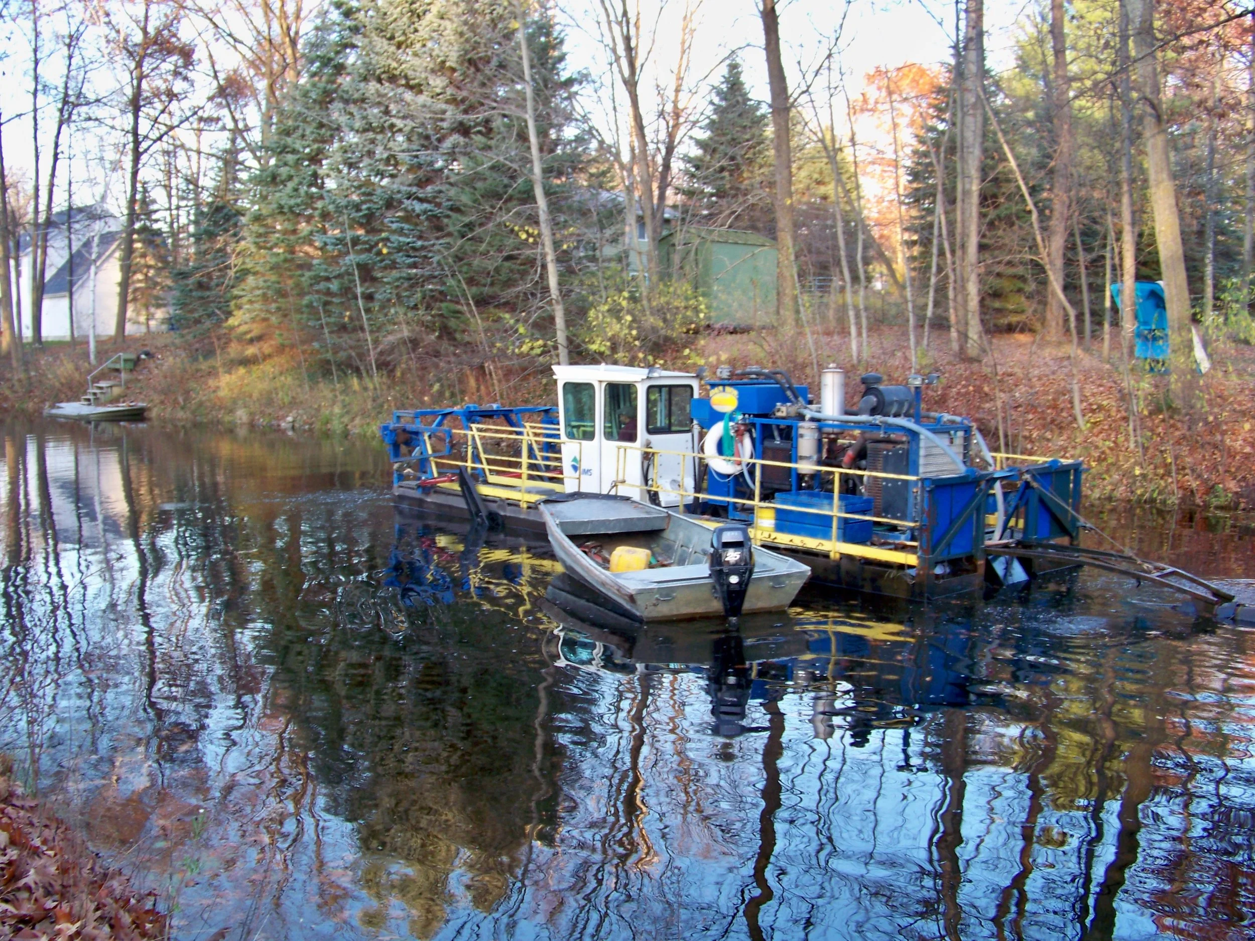 A small boat and a pontoon boat in a river with trees and houses in the background during autumn.