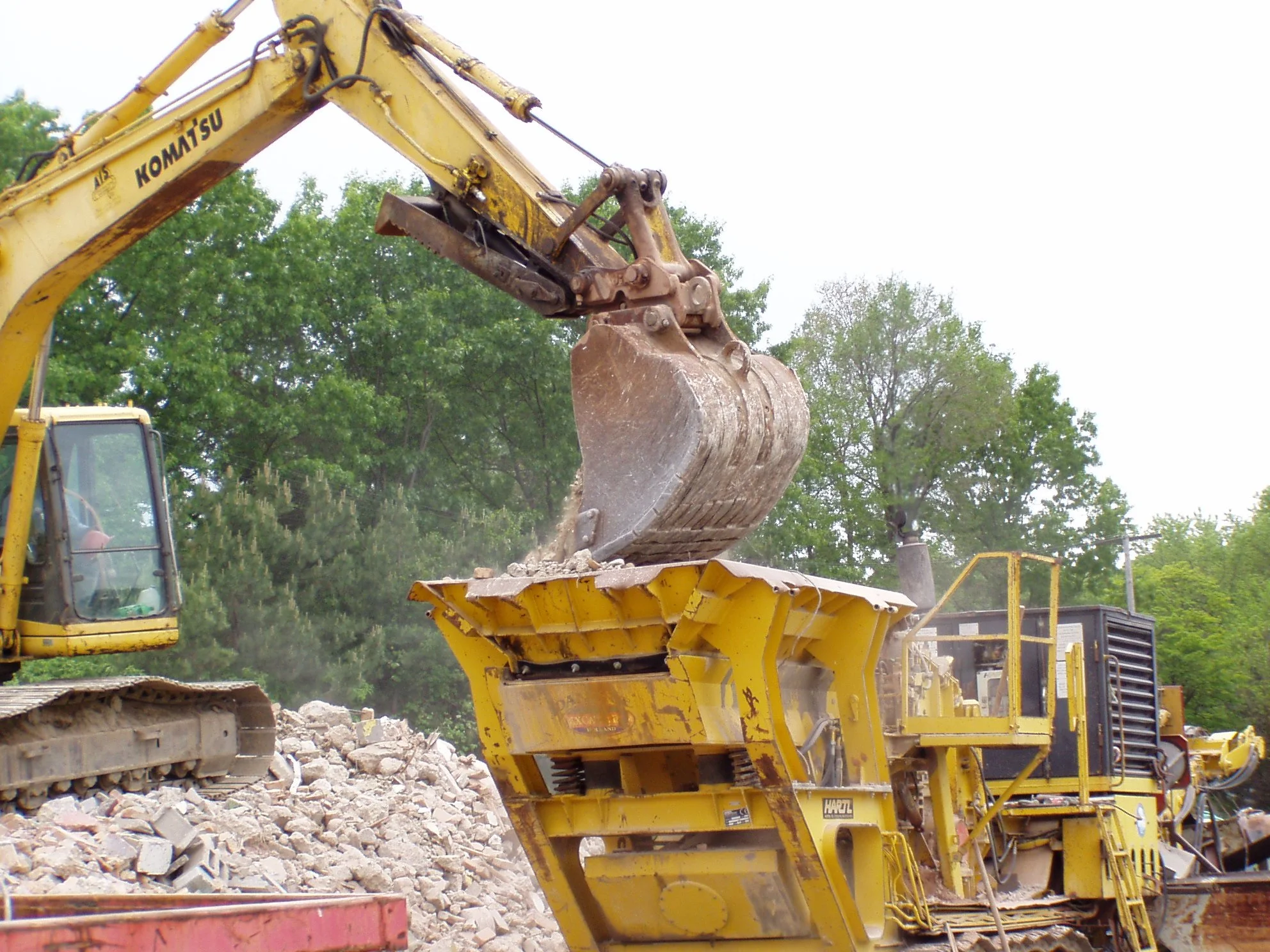 An excavator loading rocks into a large yellow rock crusher at a construction site, with green trees in the background.