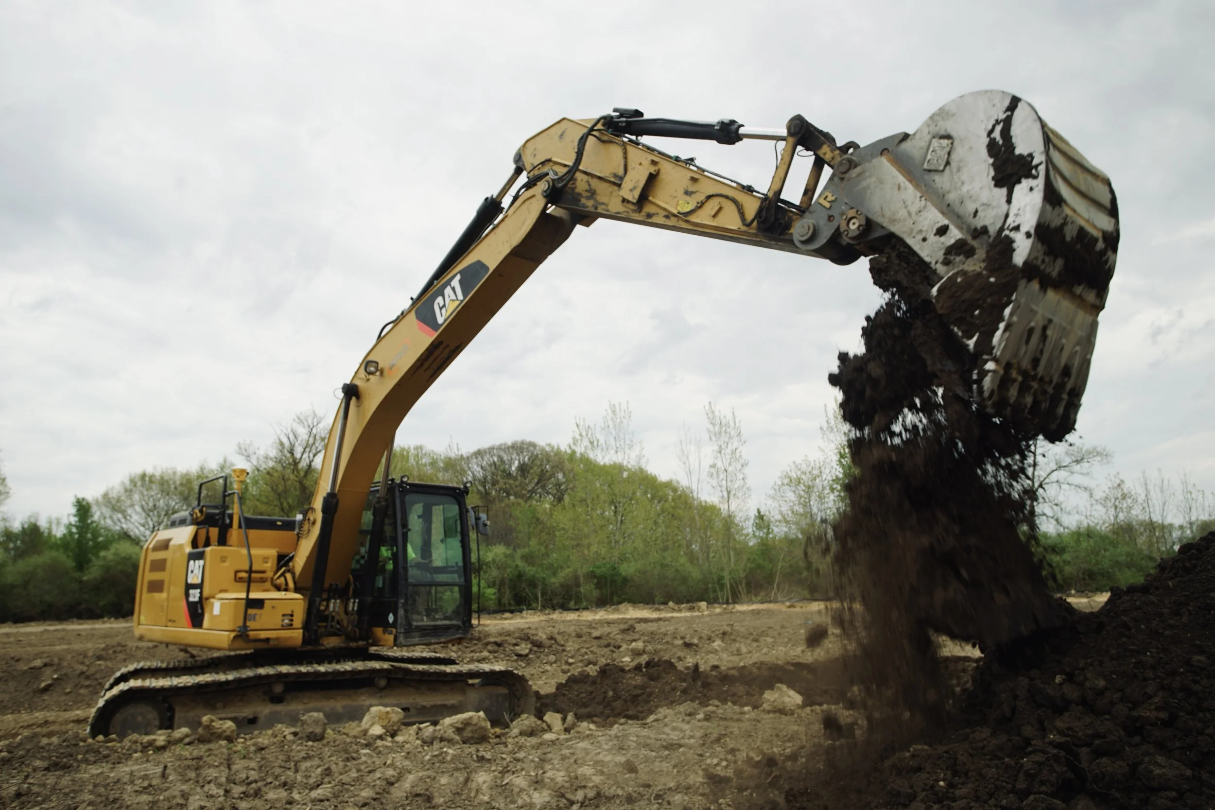 A yellow Caterpillar excavator digging and moving dark soil on a construction site with a cloudy sky and some trees in the background.