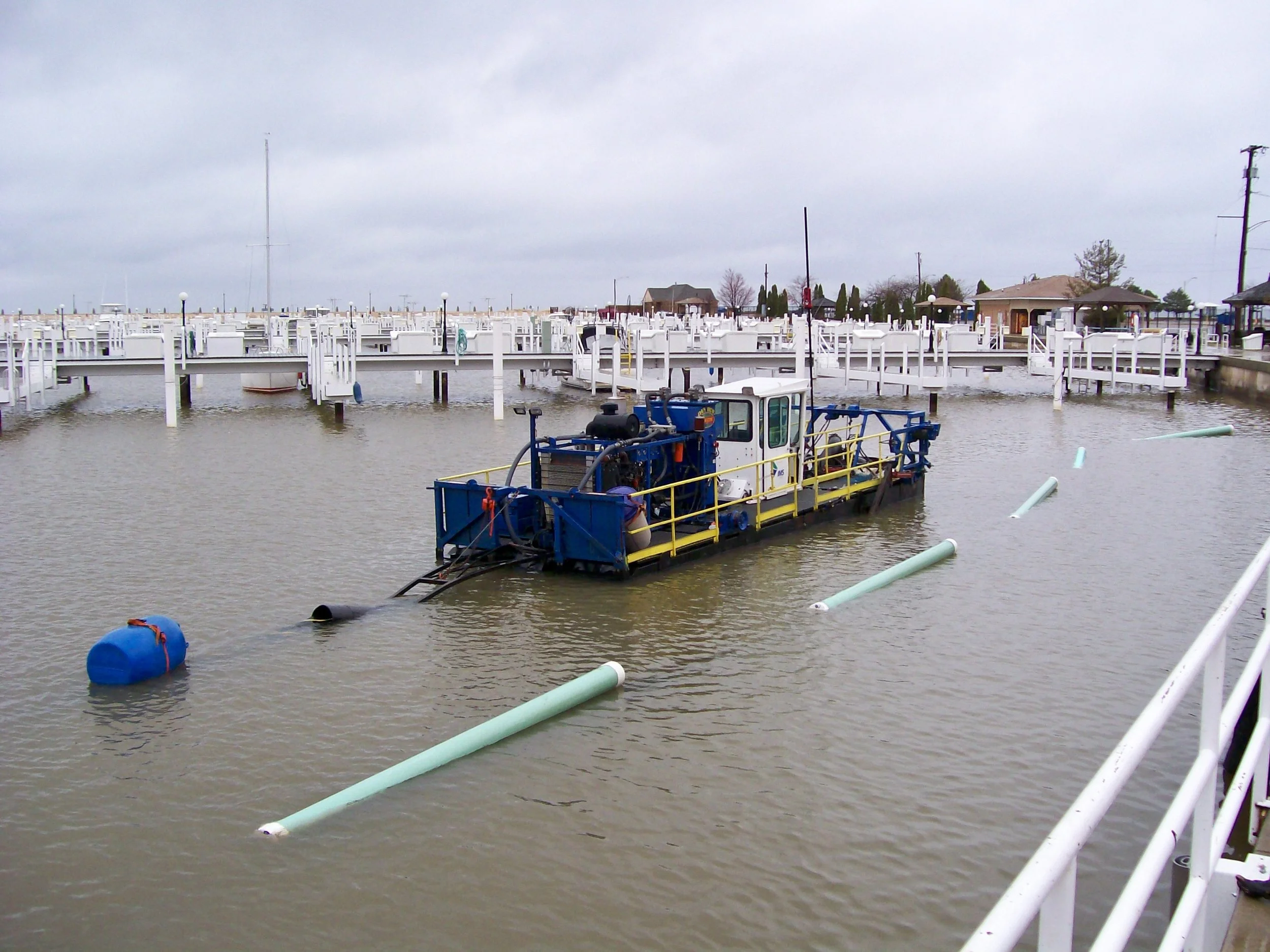 A boat cleaning or dredging machine in a marina filled with water, surrounded by floating buoy markers and white docks, with homes and overcast skies in the background.