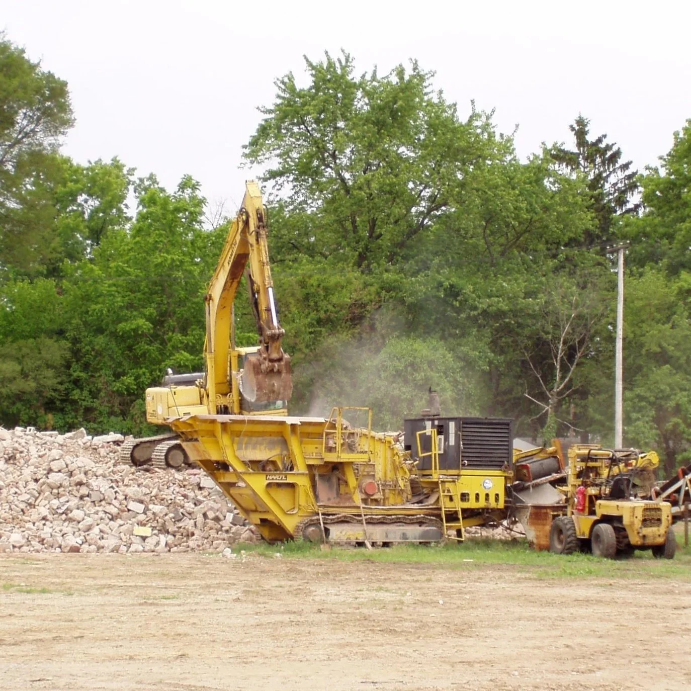 Construction site with yellow heavy machinery, including an excavator and a bulldozer, working on a pile of rocks with a background of green trees.