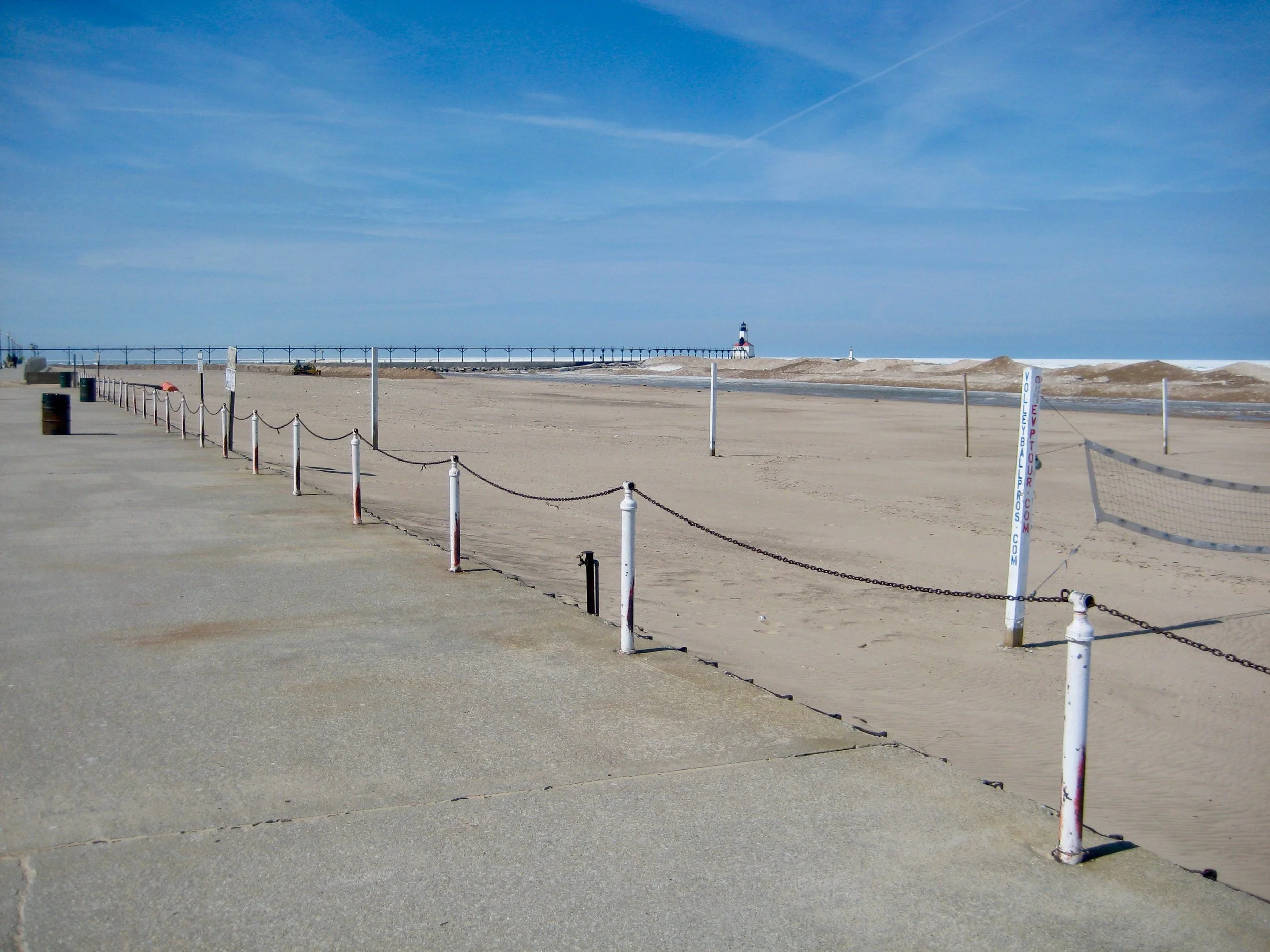 Empty sandy beach with a concrete promenade on the left and a white metal chain fence. In the distance, a lighthouse on the coast and a long pier extending into the ocean. Blue sky with a few wispy clouds.