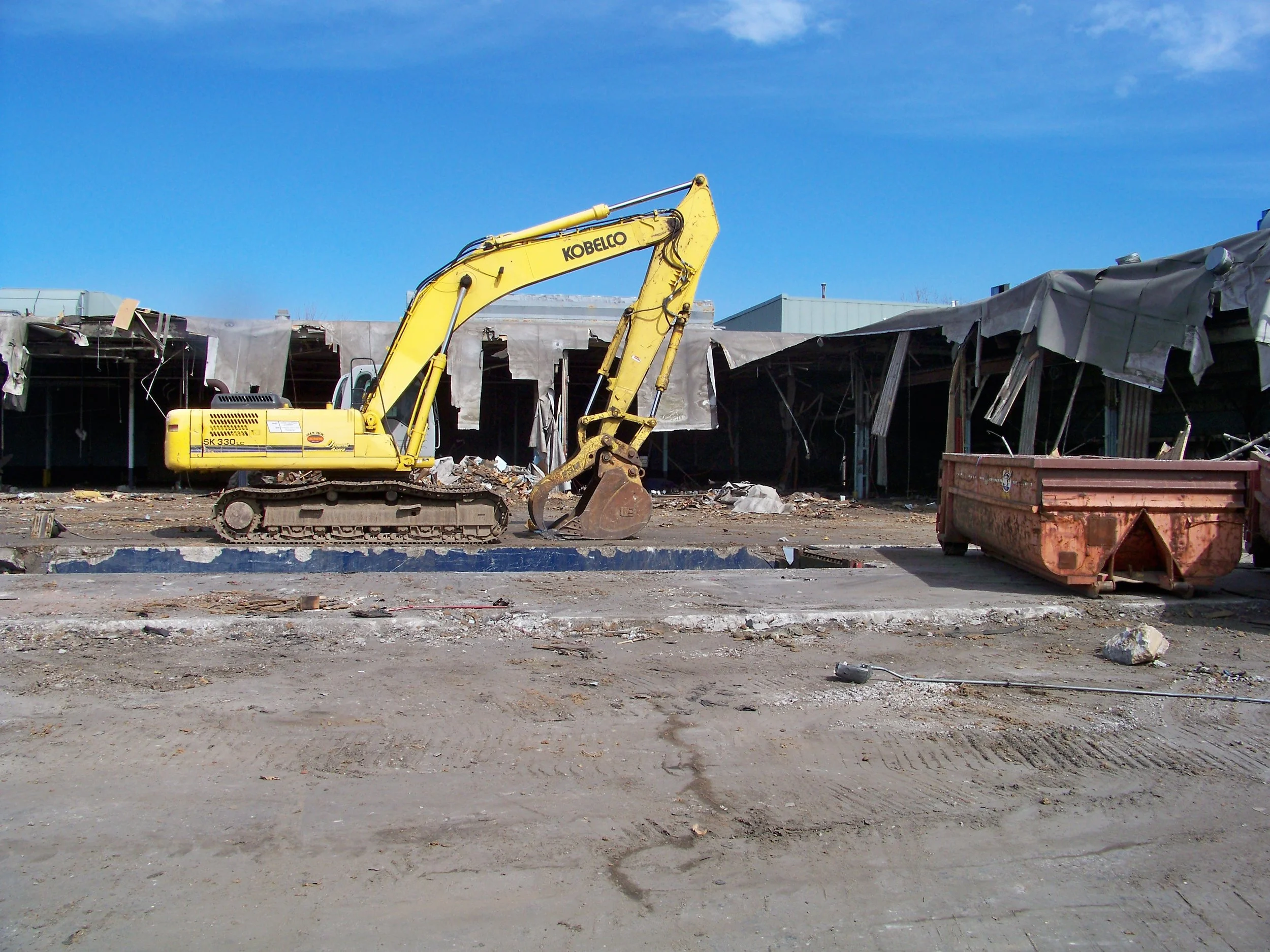 Construction site with a yellow excavator and partially demolished buildings in the background under a blue sky.