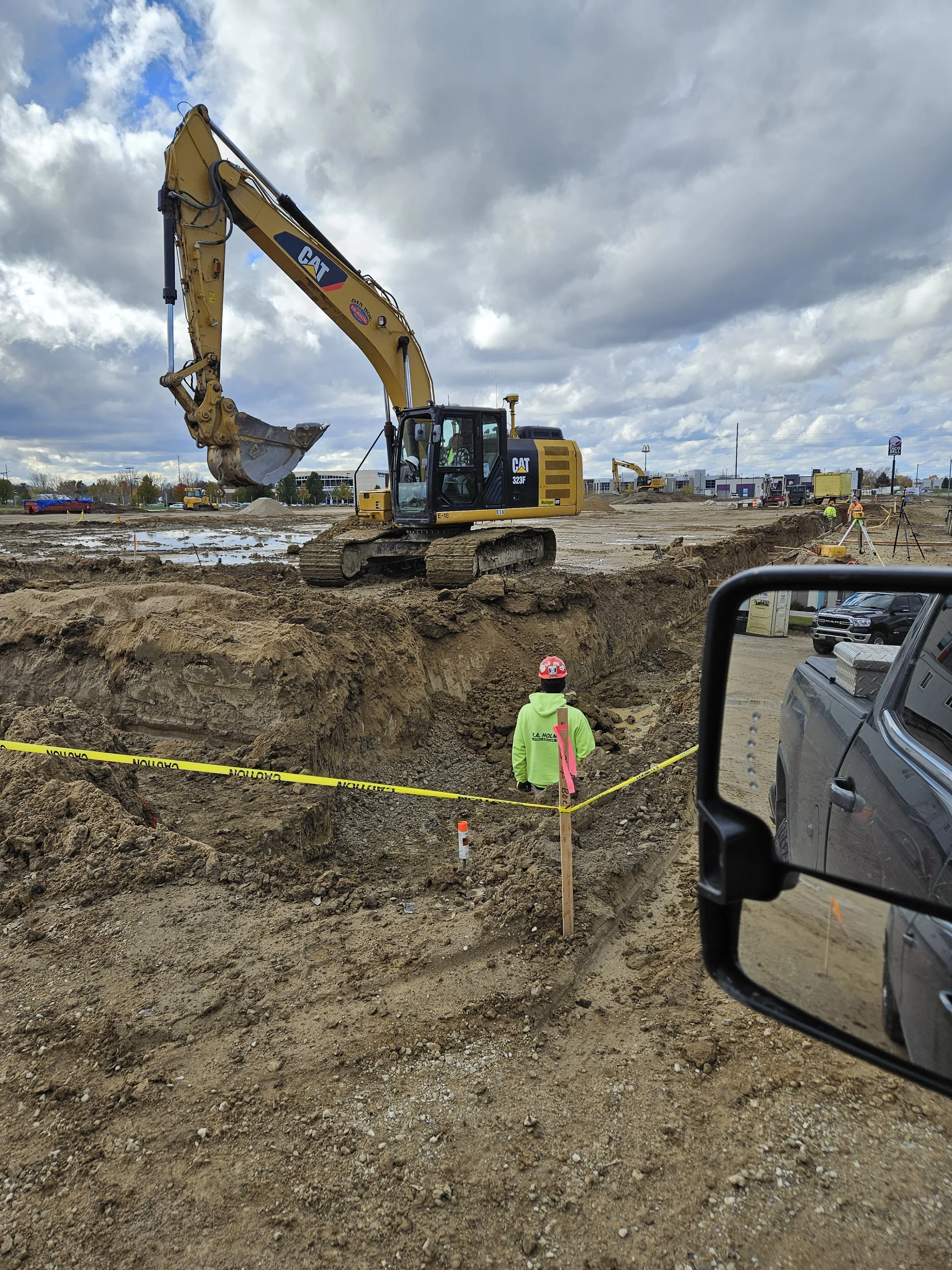 A construction site with an excavator working in the background, a worker wearing a red helmet and green jacket observing the site, and caution tape outlining the excavated area under a cloudy sky.