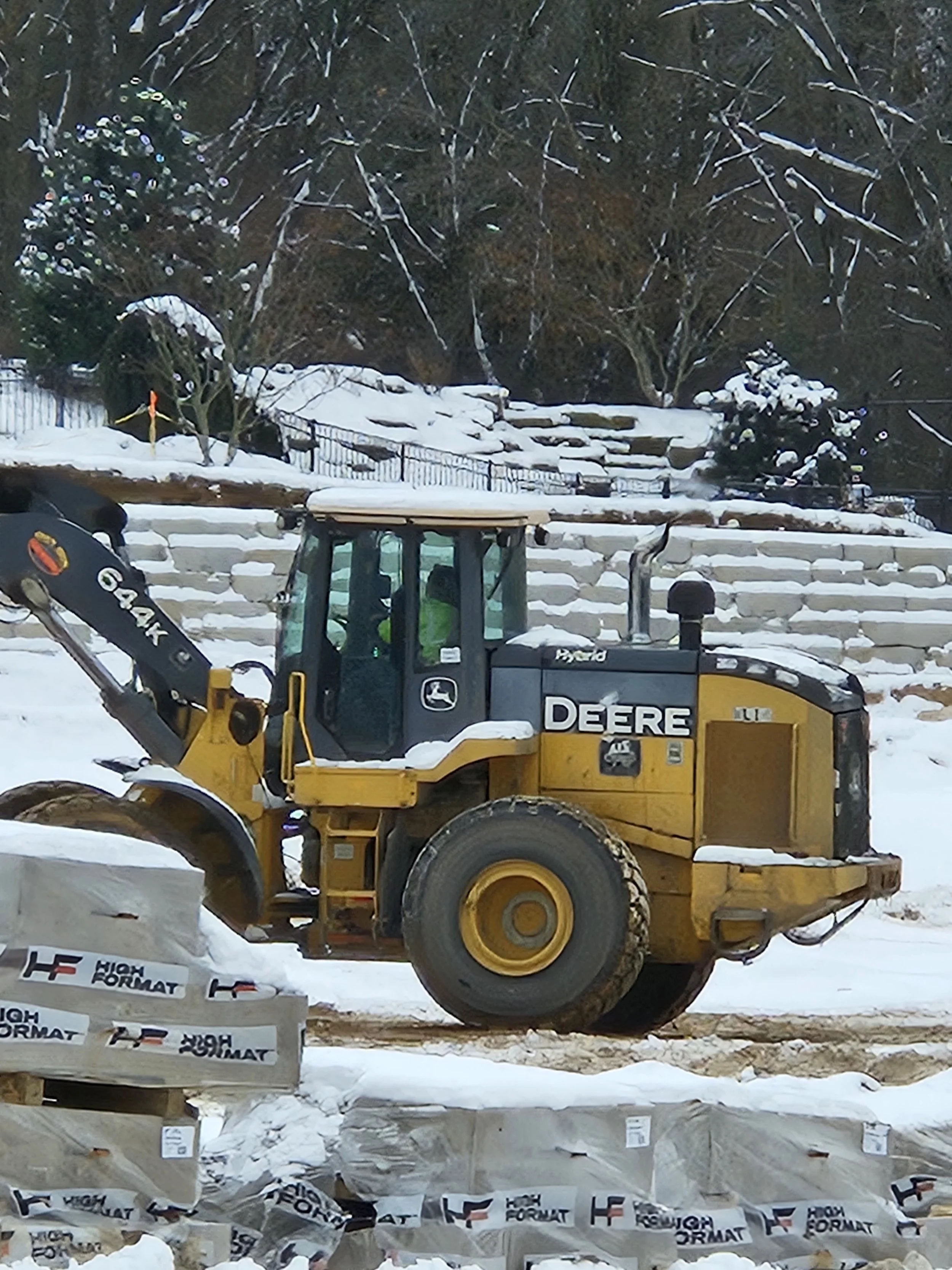 Yellow John Deere compact loader working on a snow-covered construction site in winter with building materials in foreground and snowy steps and trees in background.
