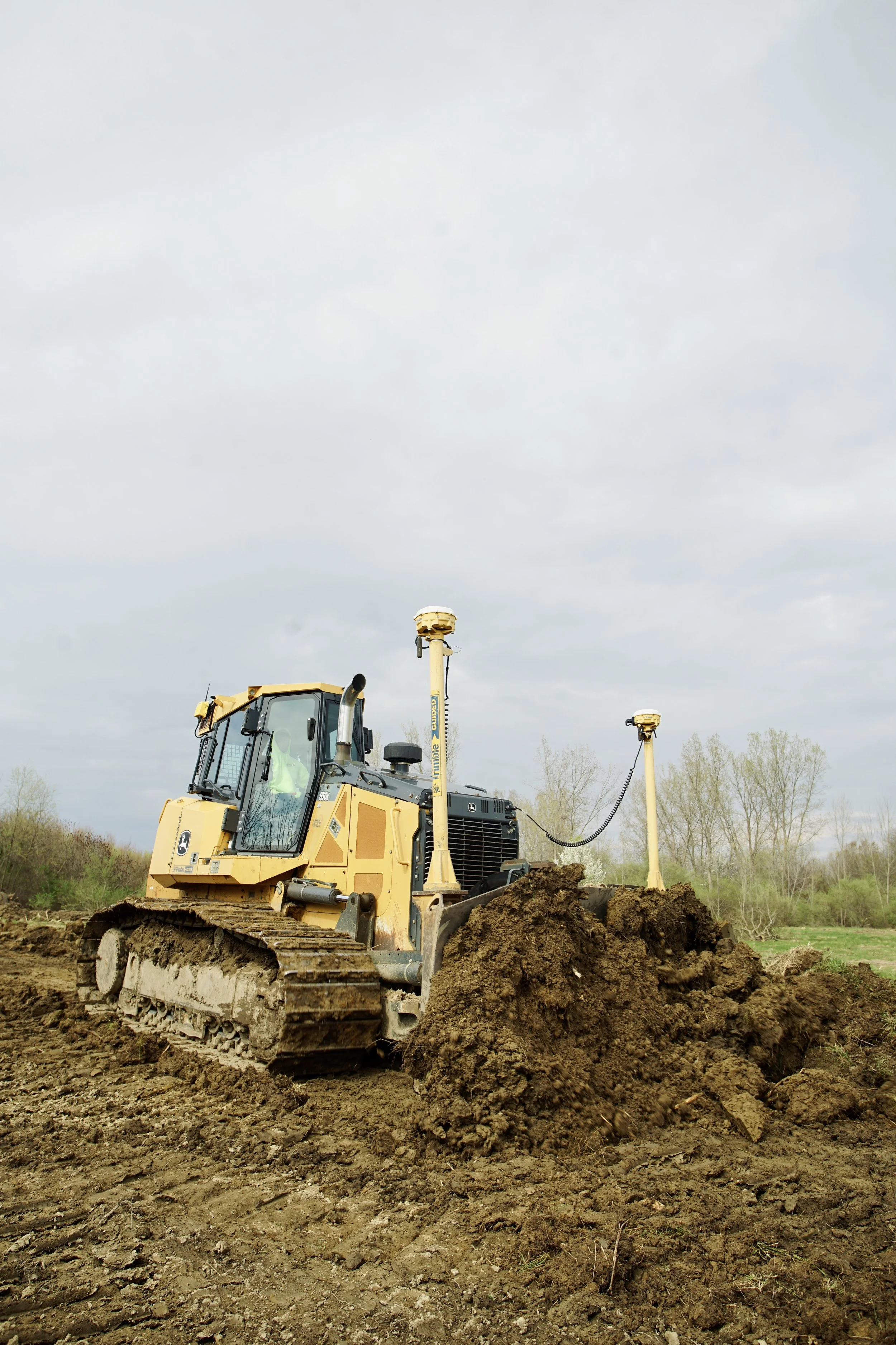 A yellow Caterpillar excavator moving dirt on a construction site with trees and cloudy sky in the background.