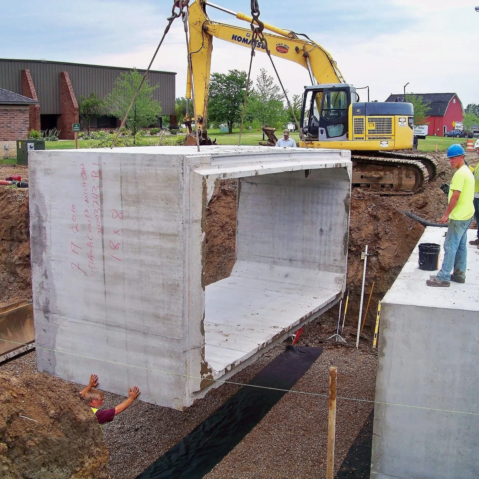 Construction workers install a large concrete pipe with a yellow excavator on a construction site.