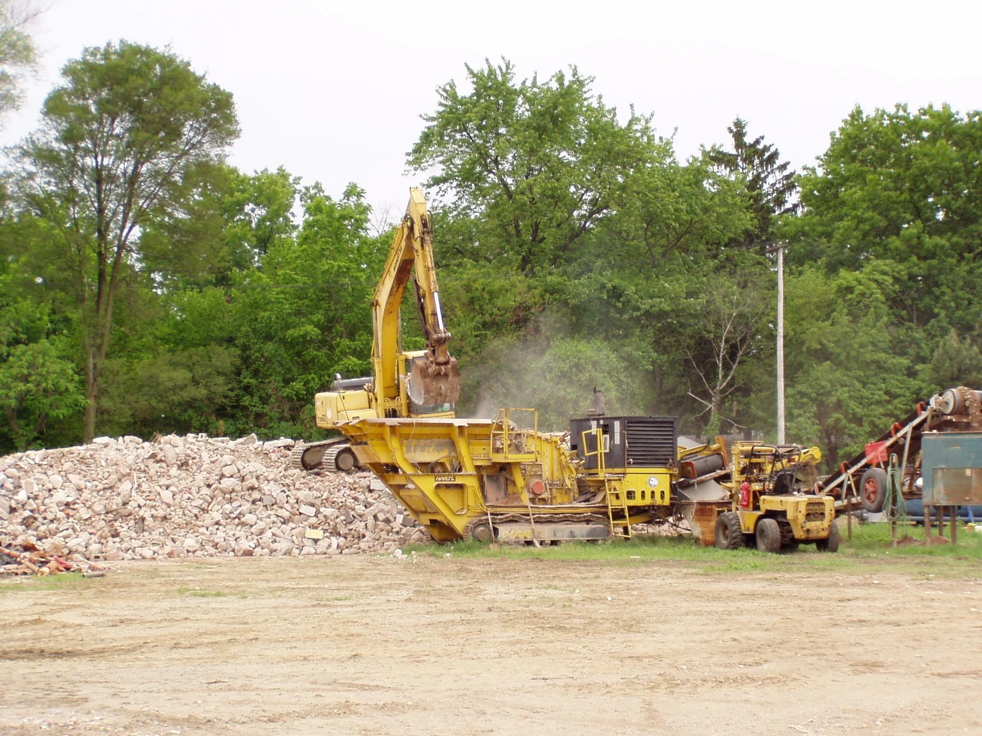 Construction site with a yellow excavator and other machinery, piles of rocks, and green trees in the background.