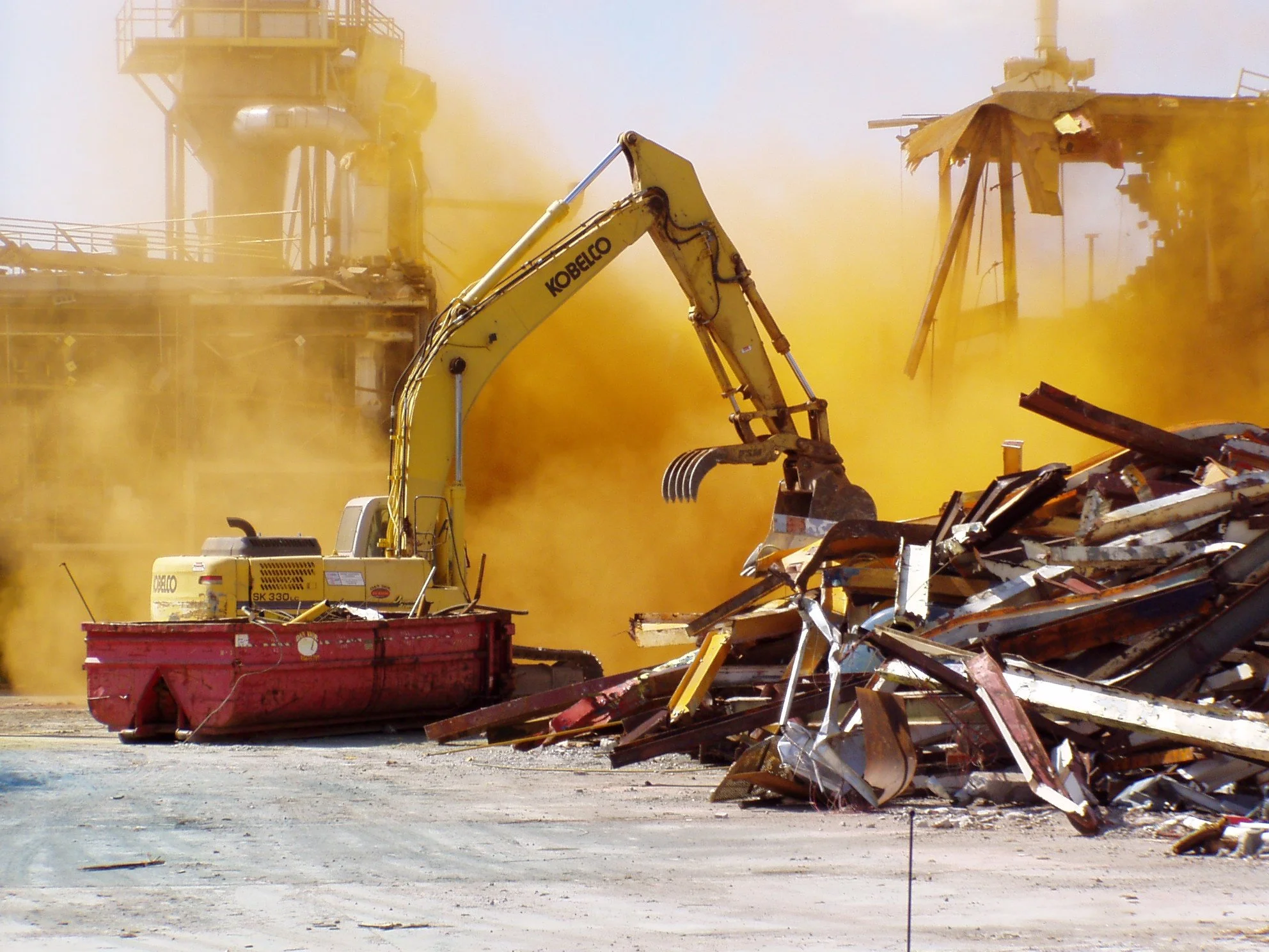 A construction excavator with the brand name Kobelco is demolishing a structure, creating a cloud of yellow dust amid a pile of rubble.
