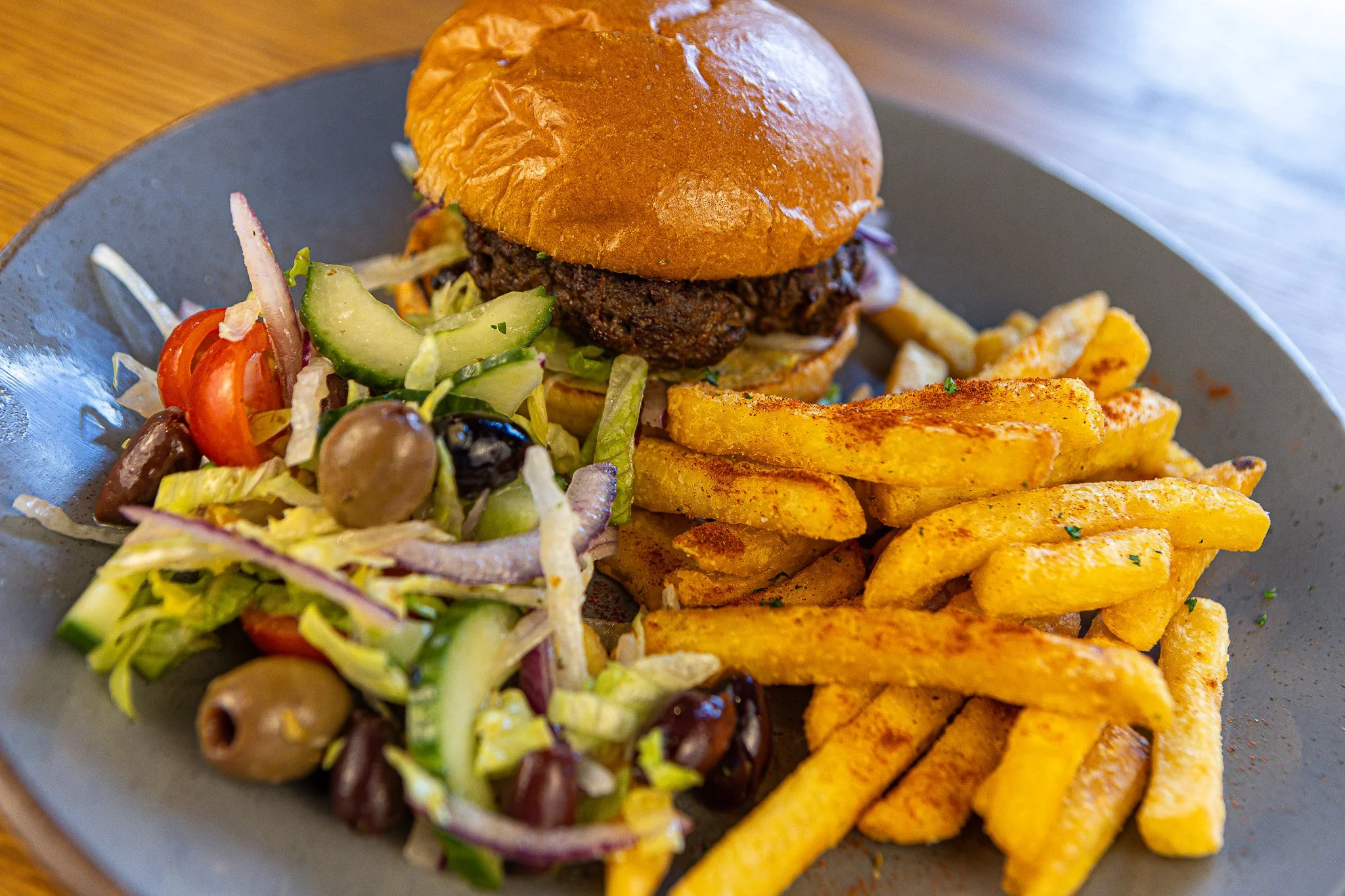 A plate of food featuring a hamburger with a bun and beef patty, alongside seasoned French fries, and a side salad with tomatoes, cucumbers, olives, and shredded lettuce.