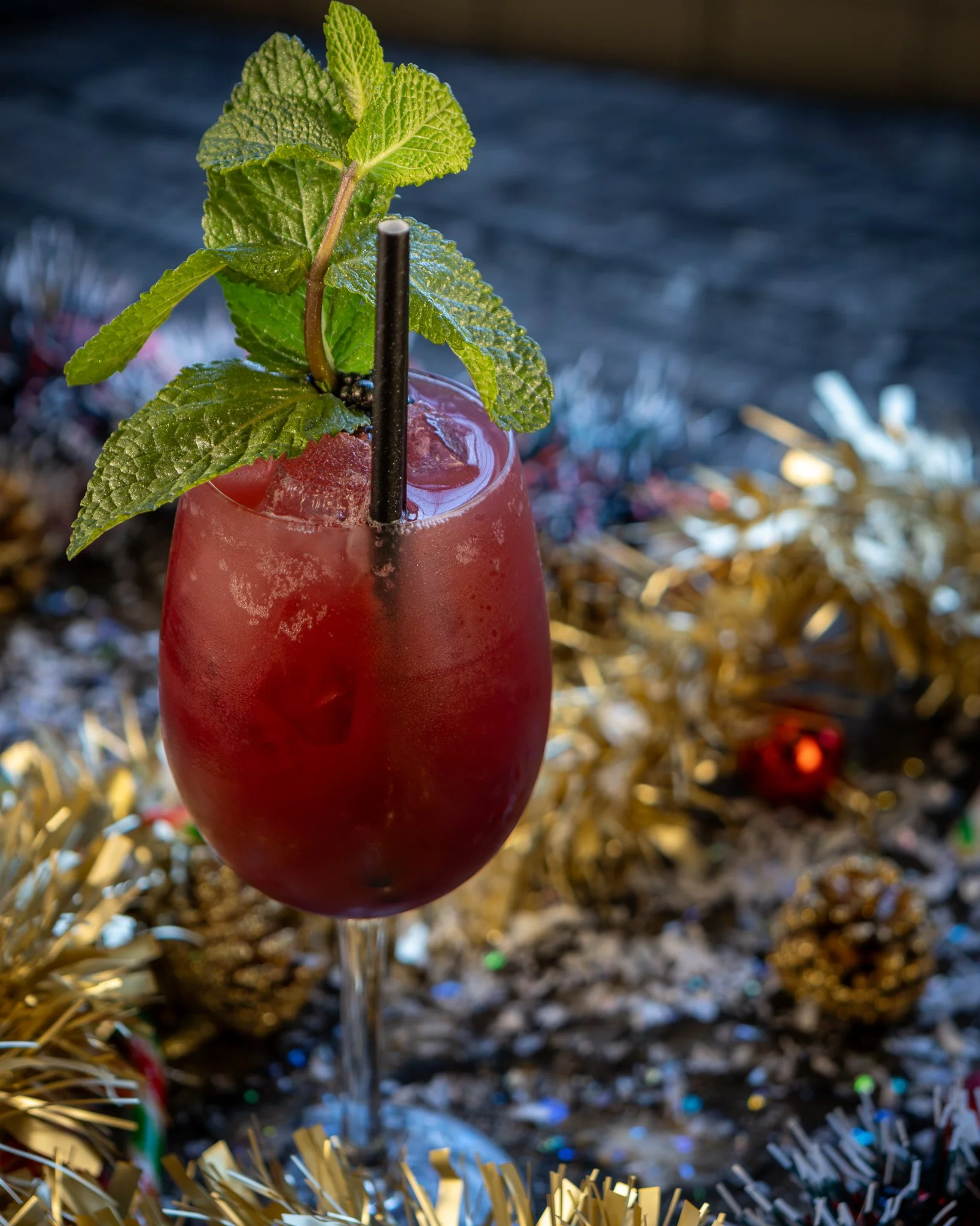 A red cocktail in a tall glass garnished with fresh mint leaves and a black straw, set against a festive, glittery background.
