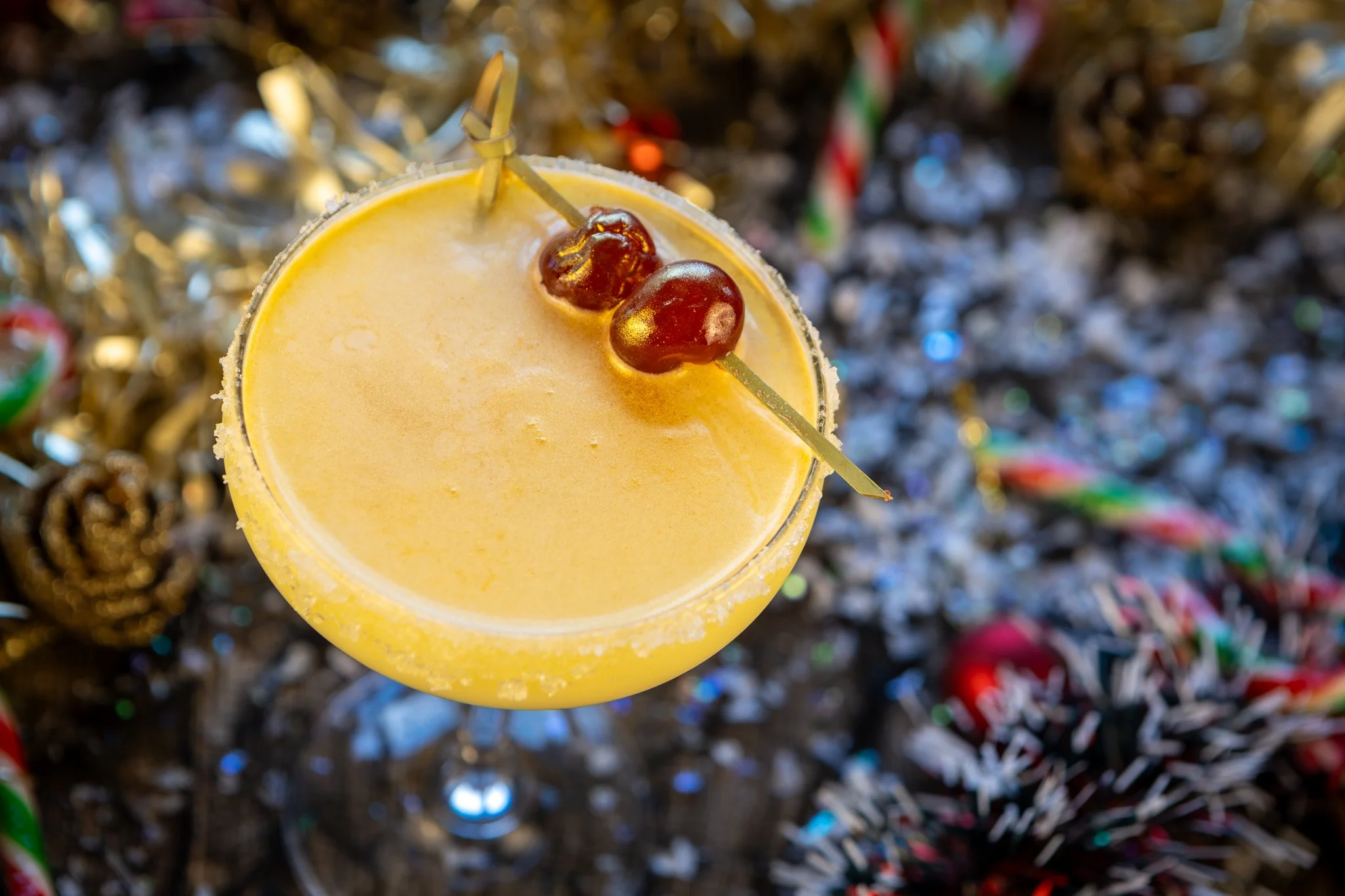 Yellow holiday cocktail garnished with two cherries on a skewer, with festive decorations in the background.