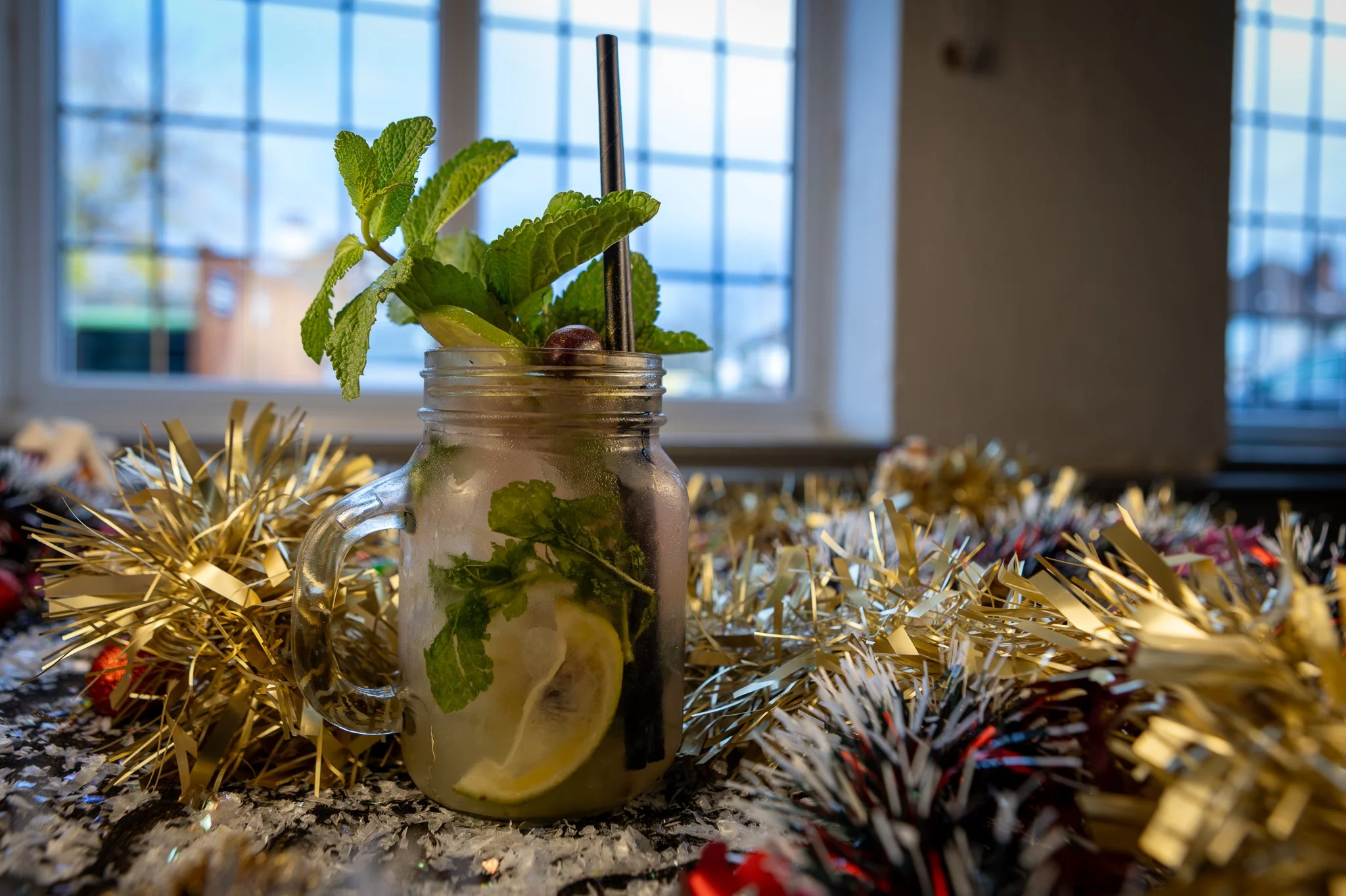 A jar filled with a mint lemonade drink with lemon slices, mint leaves, and a black straw, placed on a festive table decorated with gold tinsel.