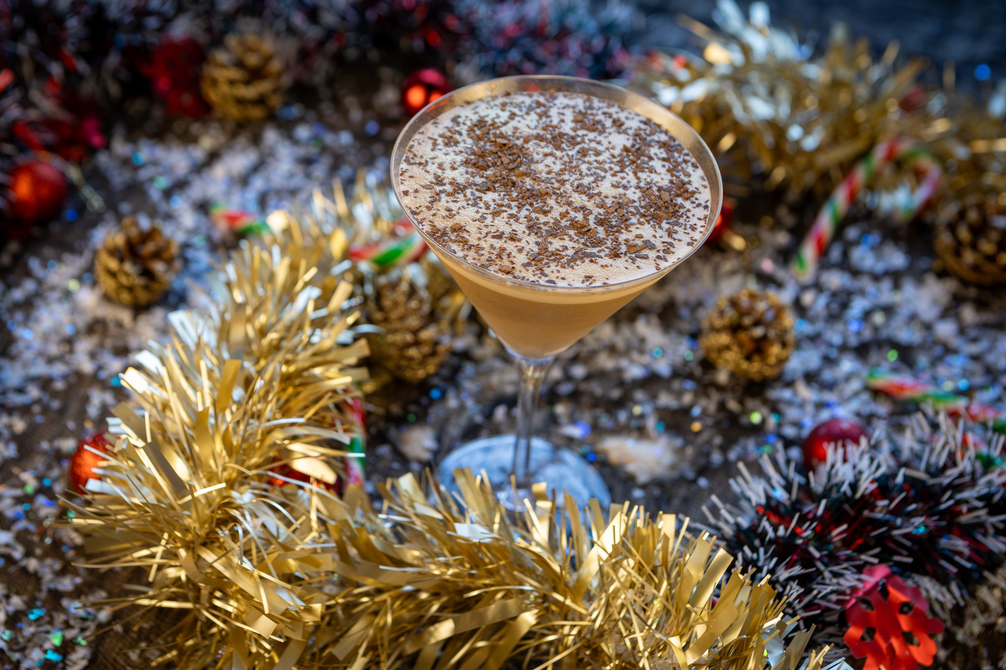 A festive holiday scene with a cocktail in a martini glass, topped with chocolate shavings, surrounded by gold tinsel, pine cones, candy canes, and Christmas ornaments.