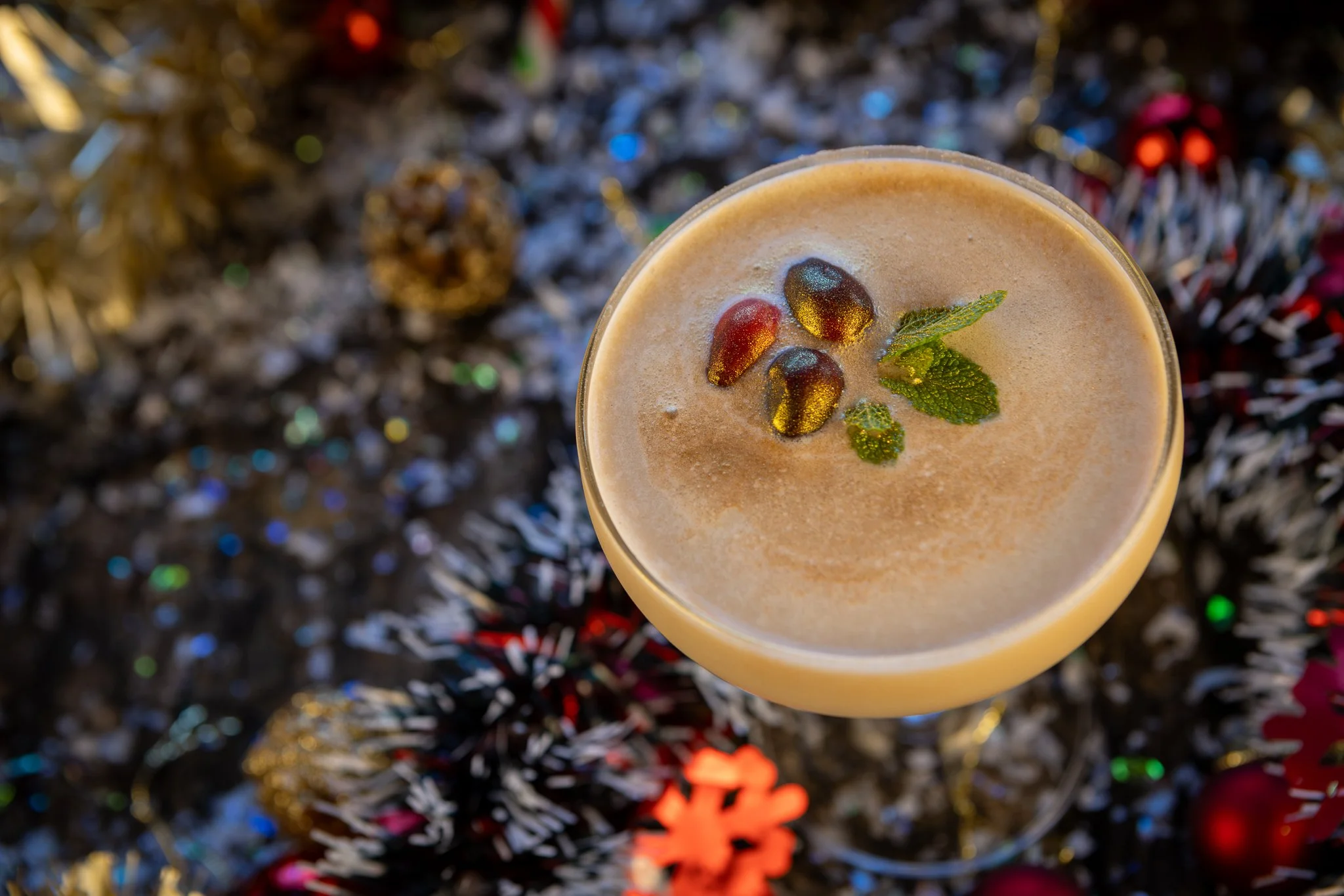 A creamy coffee drink topped with three red and black berries and fresh mint leaves, viewed from above, with colorful tinsel and holiday decorations in the background.