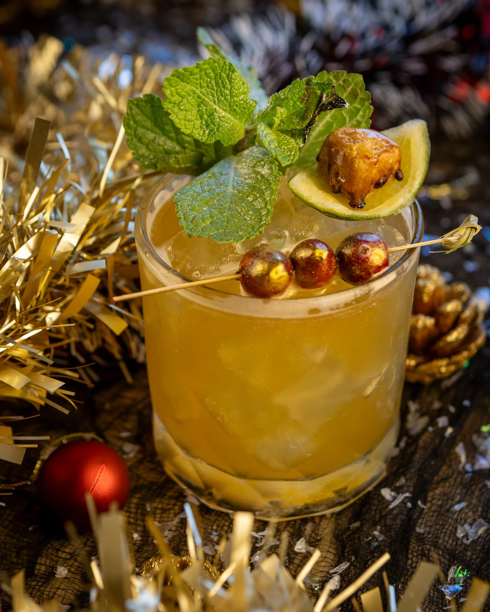 A glass of yellow cocktail garnished with fresh mint leaves, a lime wedge with a cherry on top, and skewers of red berries, surrounded by gold and red Christmas decorations and tinsel.