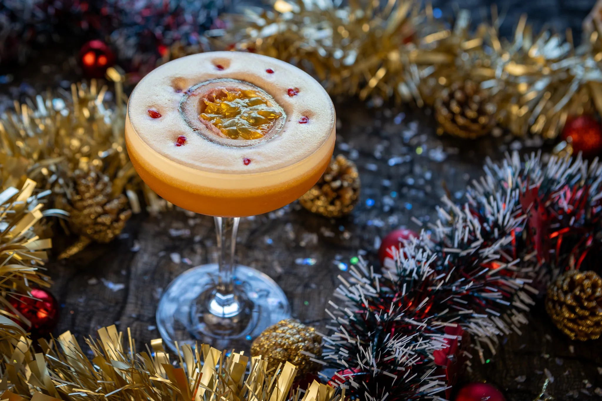 A cocktail in a coupe glass with a foam top, garnished with a passion fruit half and small red berries, surrounded by gold, red, and black holiday decorations, including tinsel, ornaments, and pinecones.