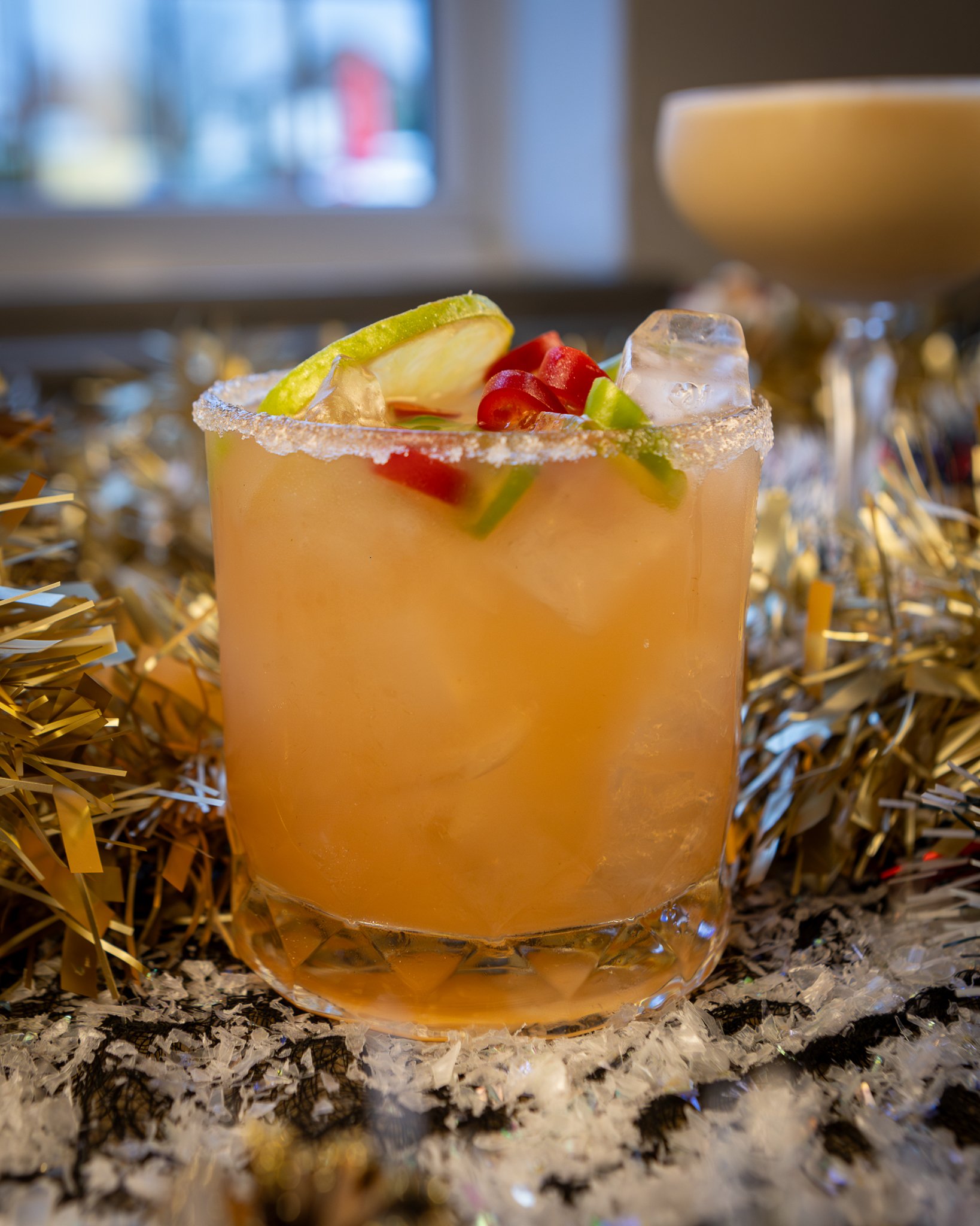 A glass of pink cocktail with lime wedge, cherry, cucumber, and ice, garnished with a salted rim, on a festive surface with gold and silver decorations.