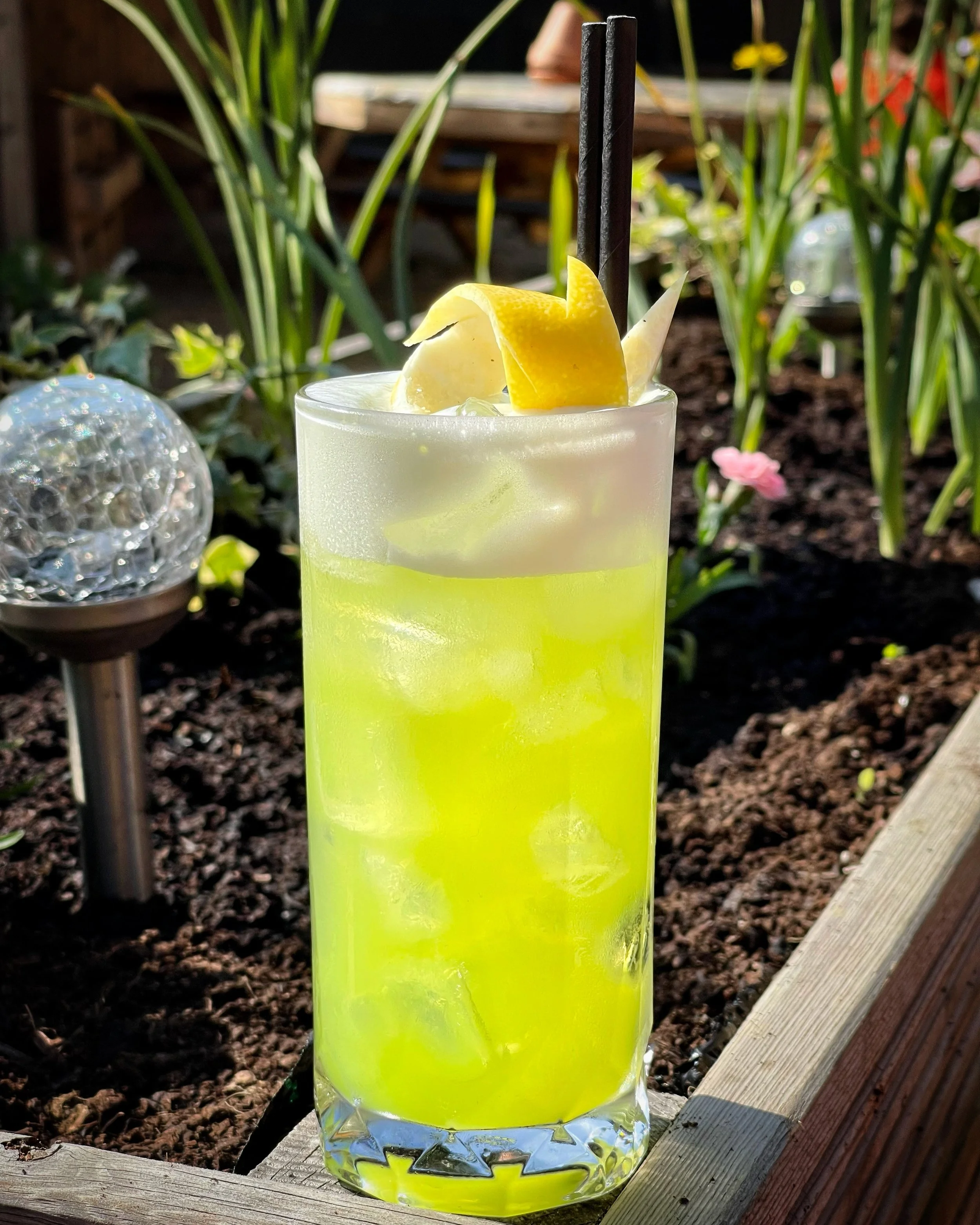 A tall glass of bright yellow lemonade with ice cubes, garnished with a lemon peel and a lemon wedge, and two black straws, set on a wooden ledge outdoors with sunlight, green plants, and garden decor in the background.