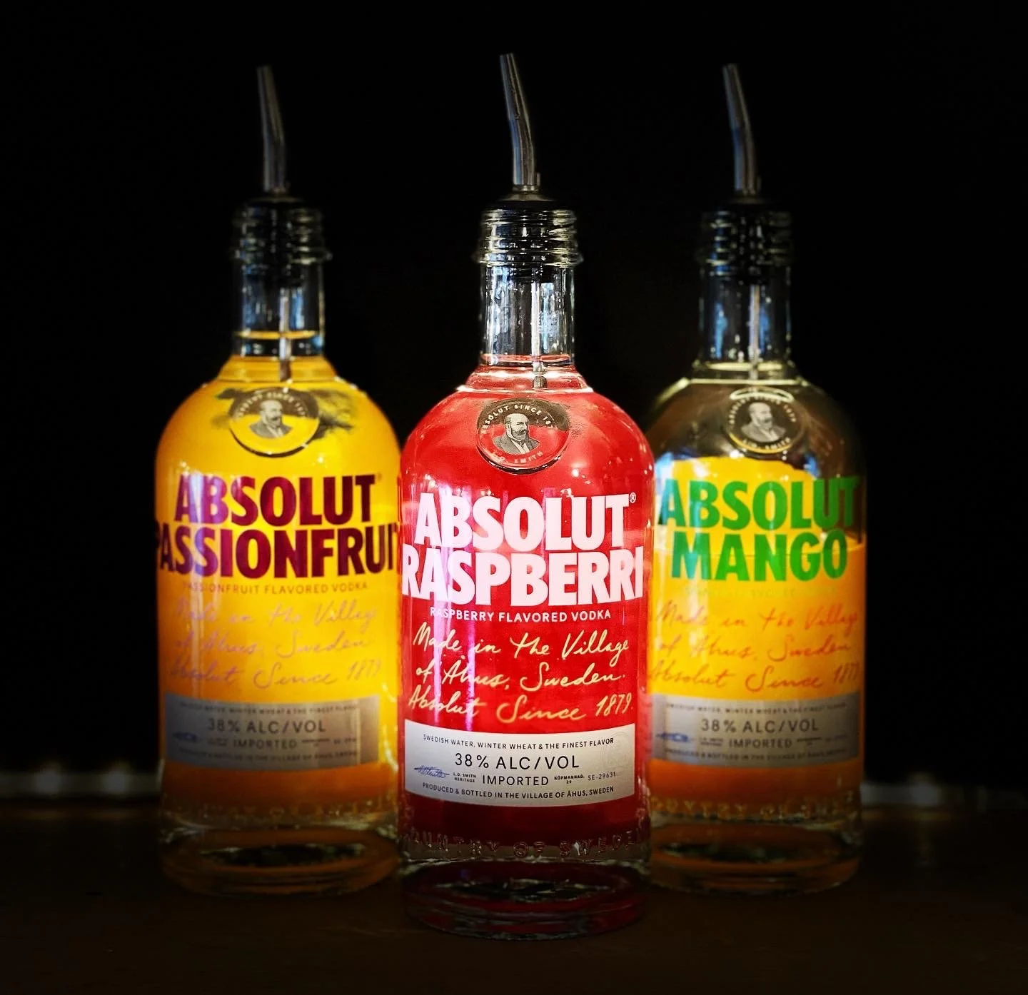 Three small bottles of Absolut flavored vodka with squirter caps, arranged in a row against a dark background. The bottles are labeled Passionfruit, Raspberry, and Mango.