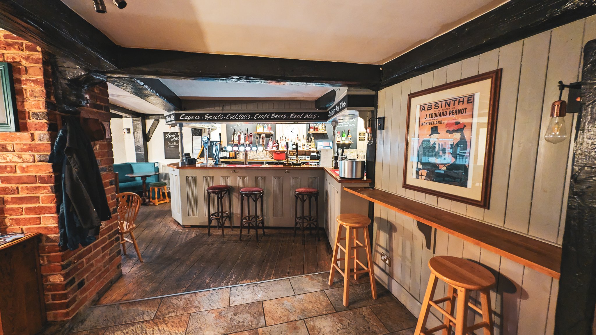 Interior of a cozy pub or bar with a wooden bar counter, bar stools, and a framed vintage-style poster on the wall. There is exposed brick, wood paneling, and warm lighting.