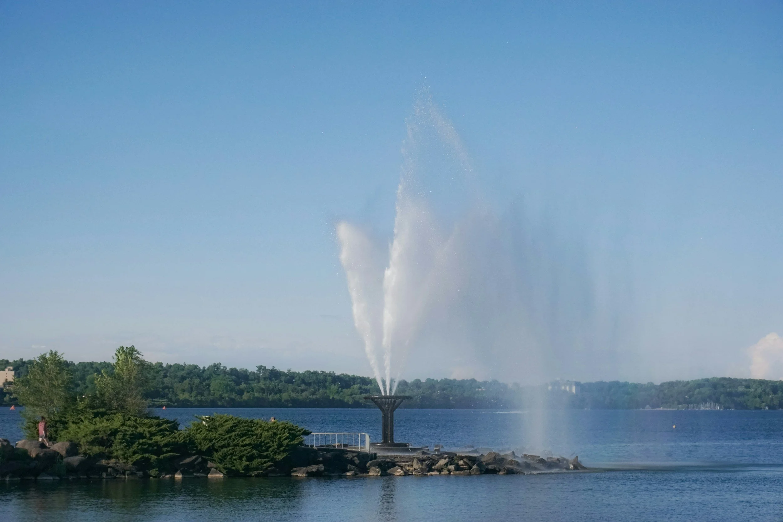 A large fountain in a lake with water spraying upwards, surrounded by trees and people enjoying the area, with a clear blue sky overhead.