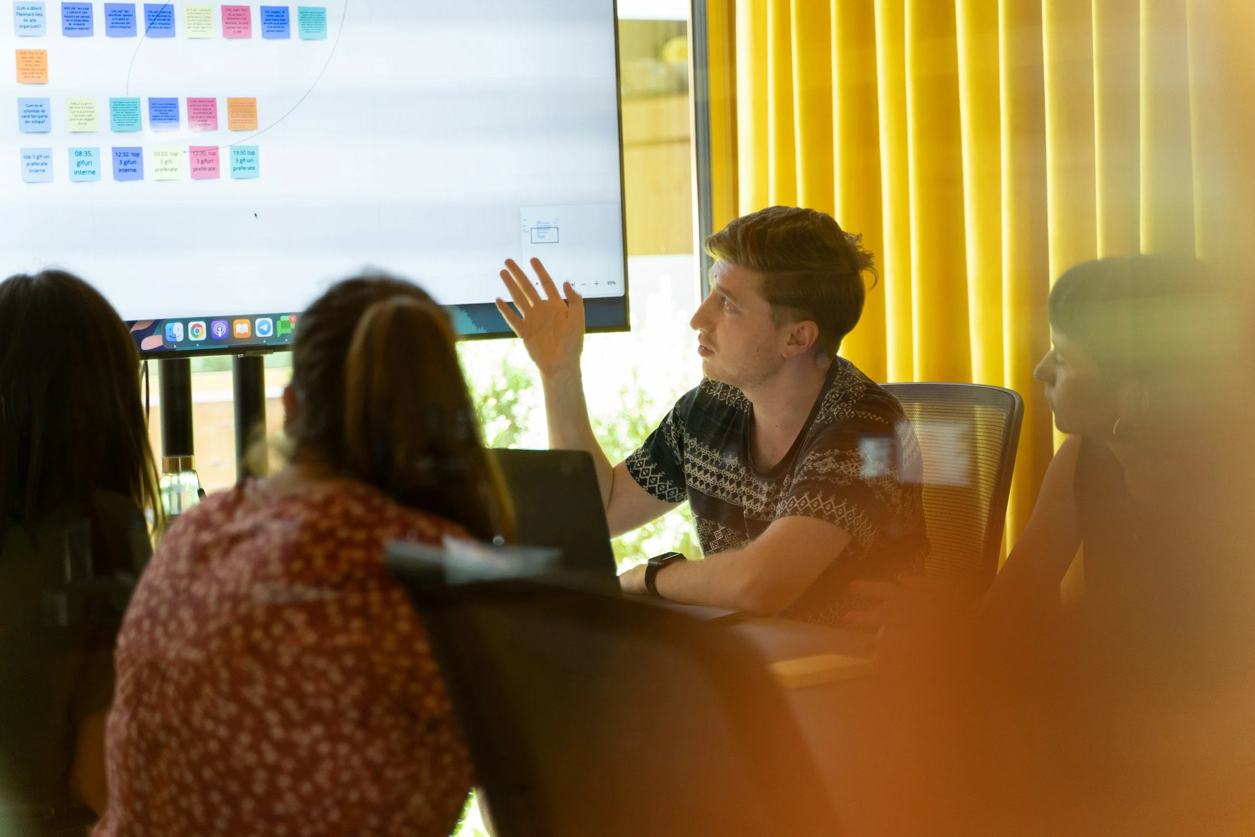 Meeting with four people, one man presenting with a large screen displaying colorful sticky notes, in a room with yellow curtains, sunlight visible outside.
