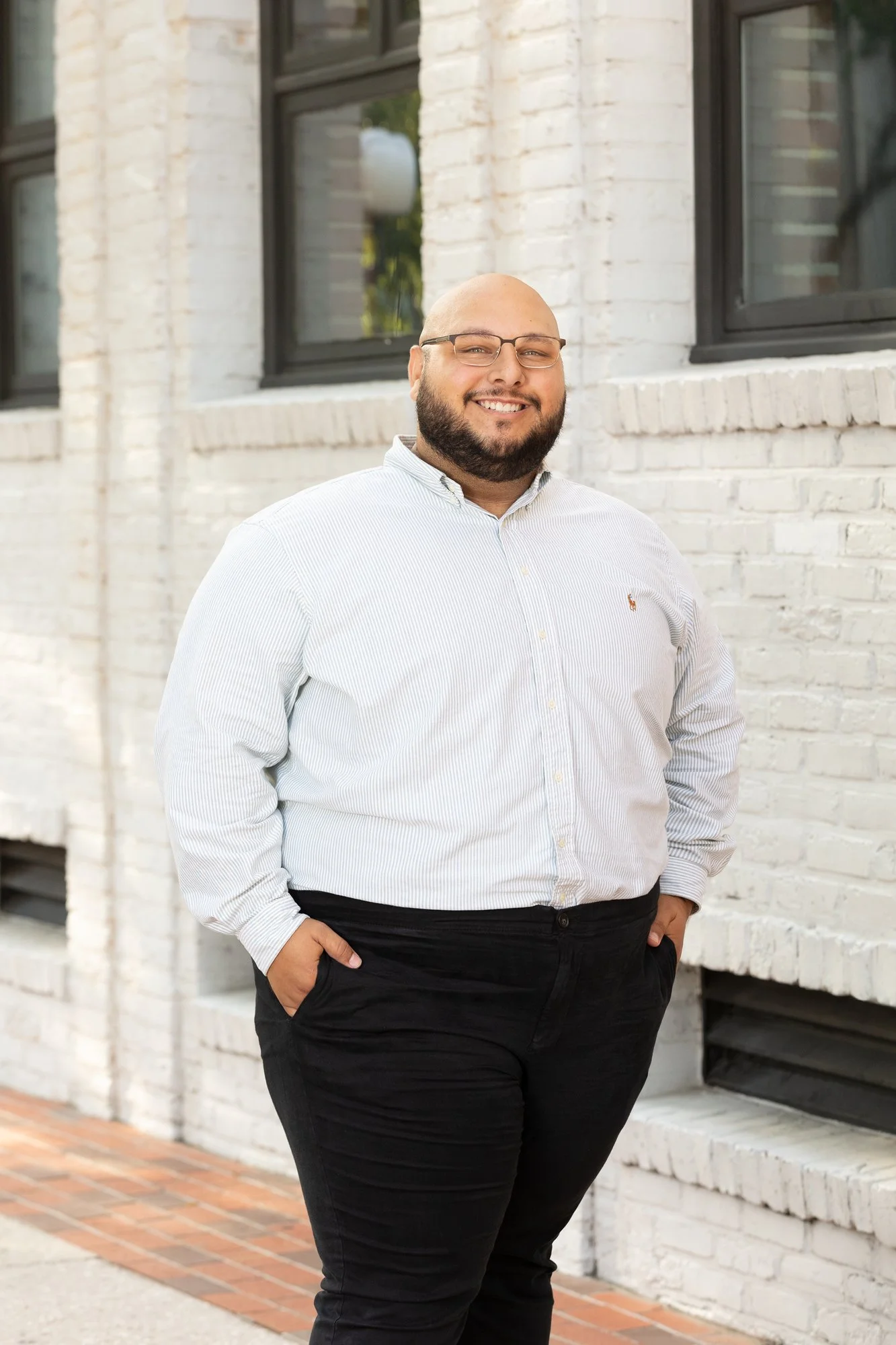 A smiling man with a beard, glasses, and a shaved head standing outdoors in front of a white brick building, wearing a white and blue striped button-up shirt and black pants.