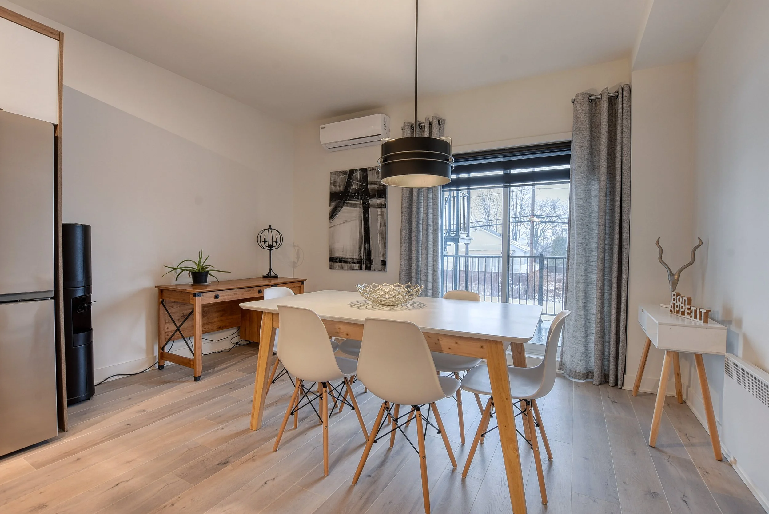 Salle à manger moderne avec table en bois clair, six chaises blanches, grand balcon, fenêtres avec rideaux gris, décor minimaliste, plantes vertes, œuvre artistique en noir et blanc, lampadaire noir, corner décoratif avec un motif en bois et la phras
