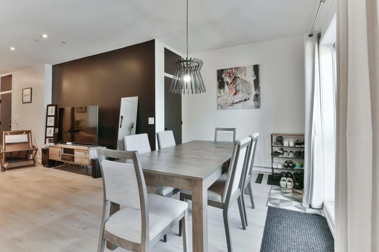 Salle à manger avec une table en bois entourée de six chaises, une petite étagère à chaussures, une porte-fenêtre avec rideaux, un tableau abstrait sur le mur blanc, et un luminaire moderne suspendu au plafond.