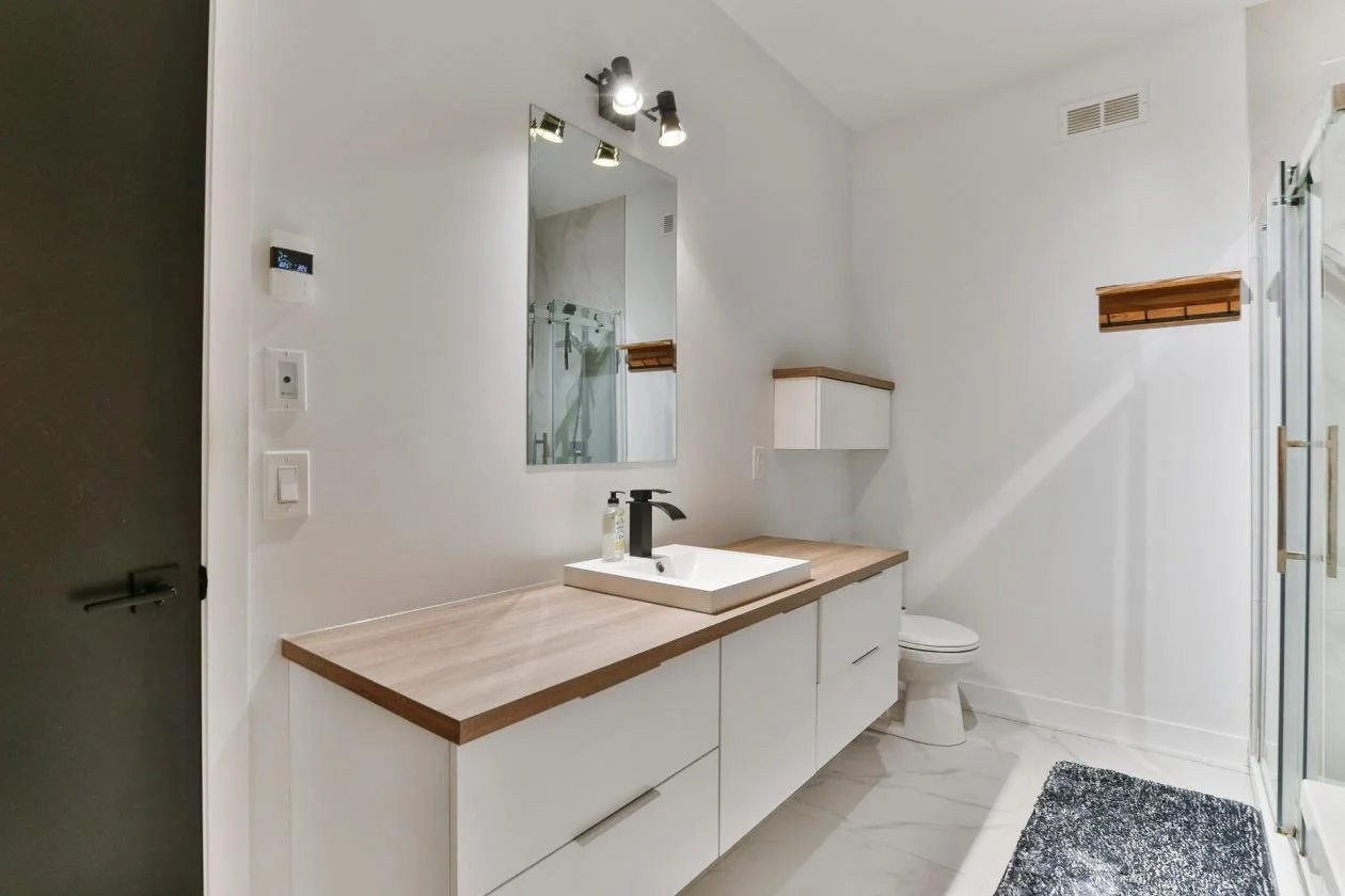Salle de bain moderne avec miroir, lavabo blanc, meuble en bois, toilette, porte en verre, tapis noir et blanc, murs blancs et éclairage encastré.