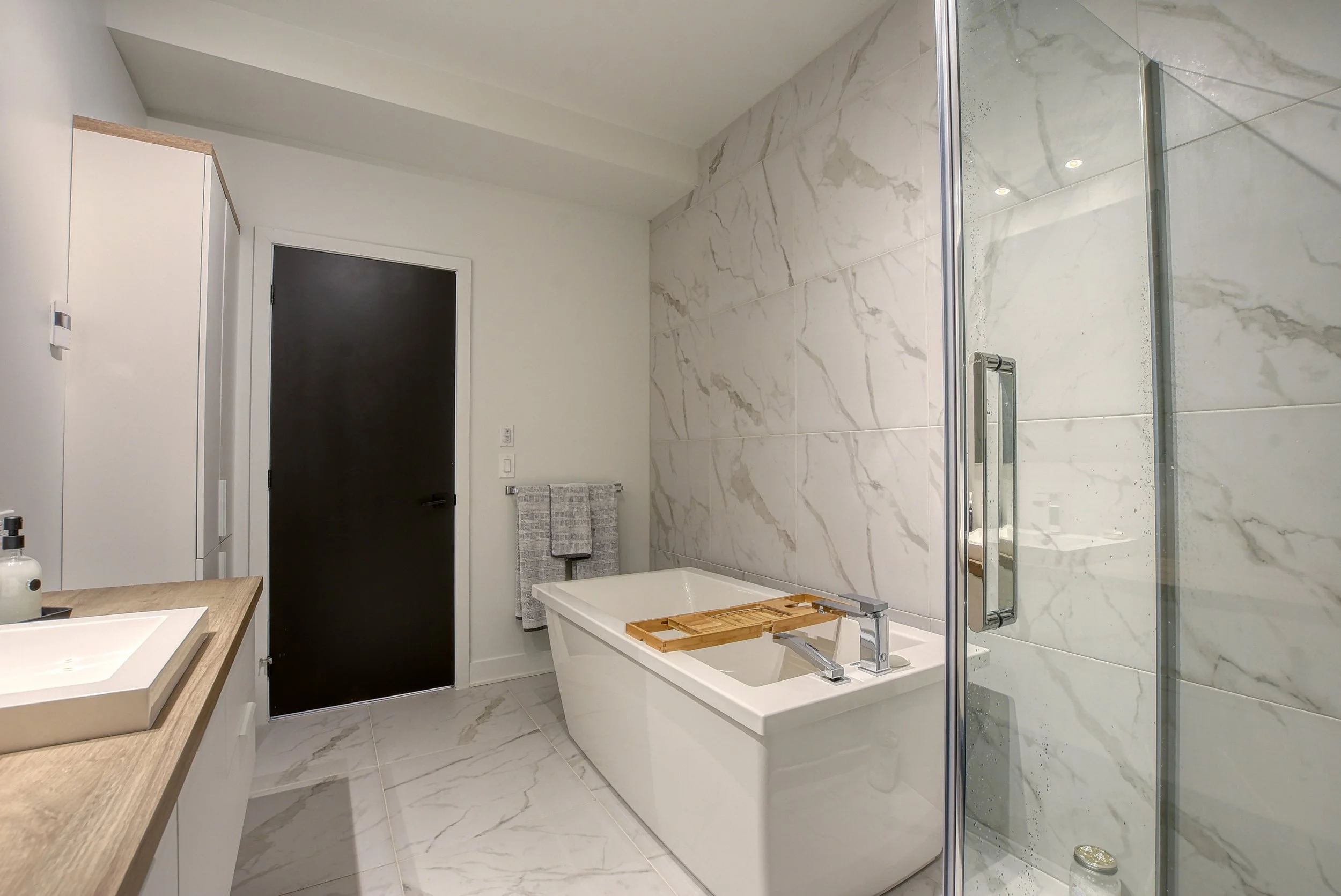 Salle de bain moderne avec baignoire, douche en verre, lavabo sur plan en bois, murs et sol en marbre blanc avec veines grises, porte noire, serviettes grises suspendues.