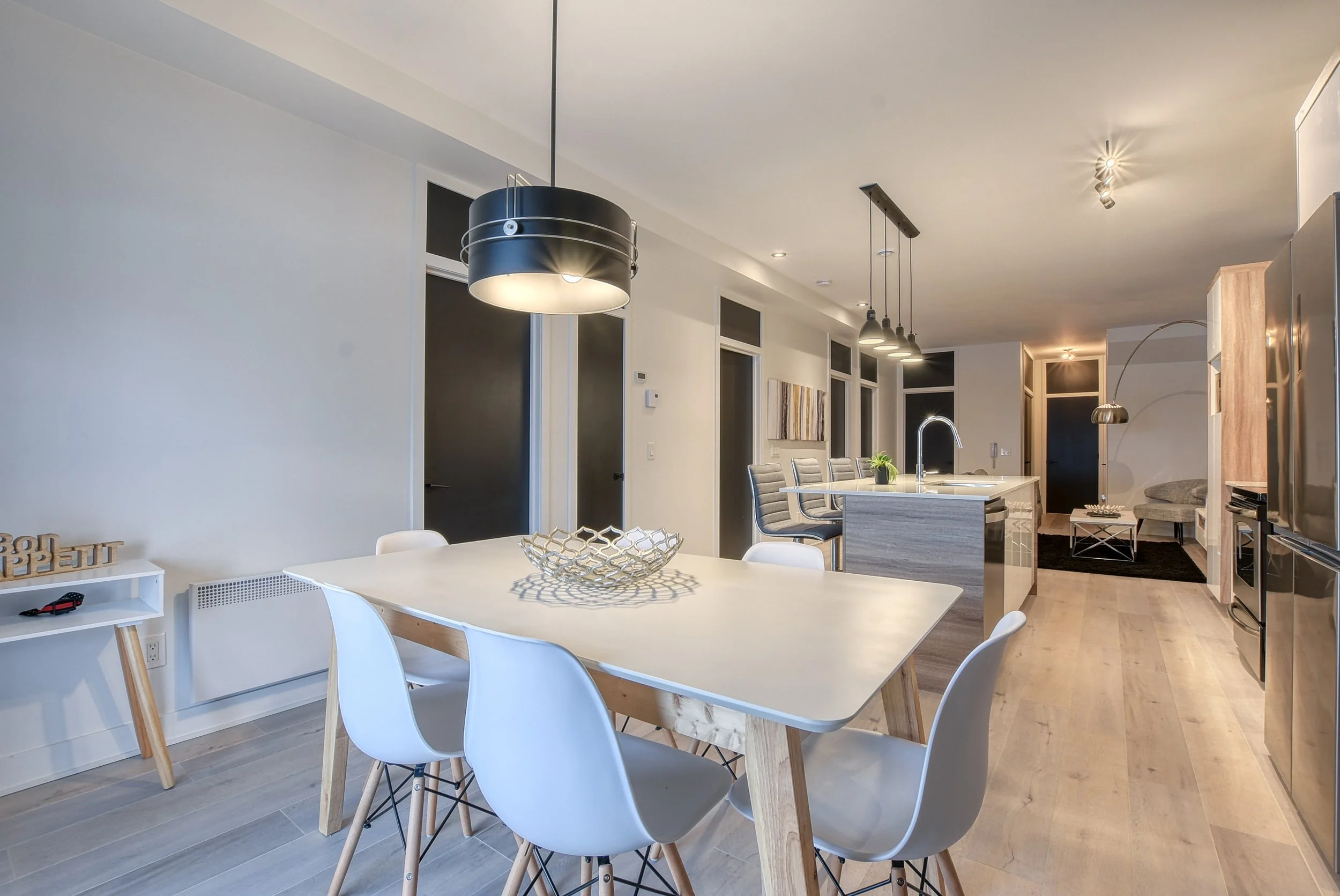 Intérieur moderne d'une cuisine ouverte avec une table à manger blanche, six chaises blanches, un îlot de cuisine avec évier, et un coin salon avec fauteuils et tapis.