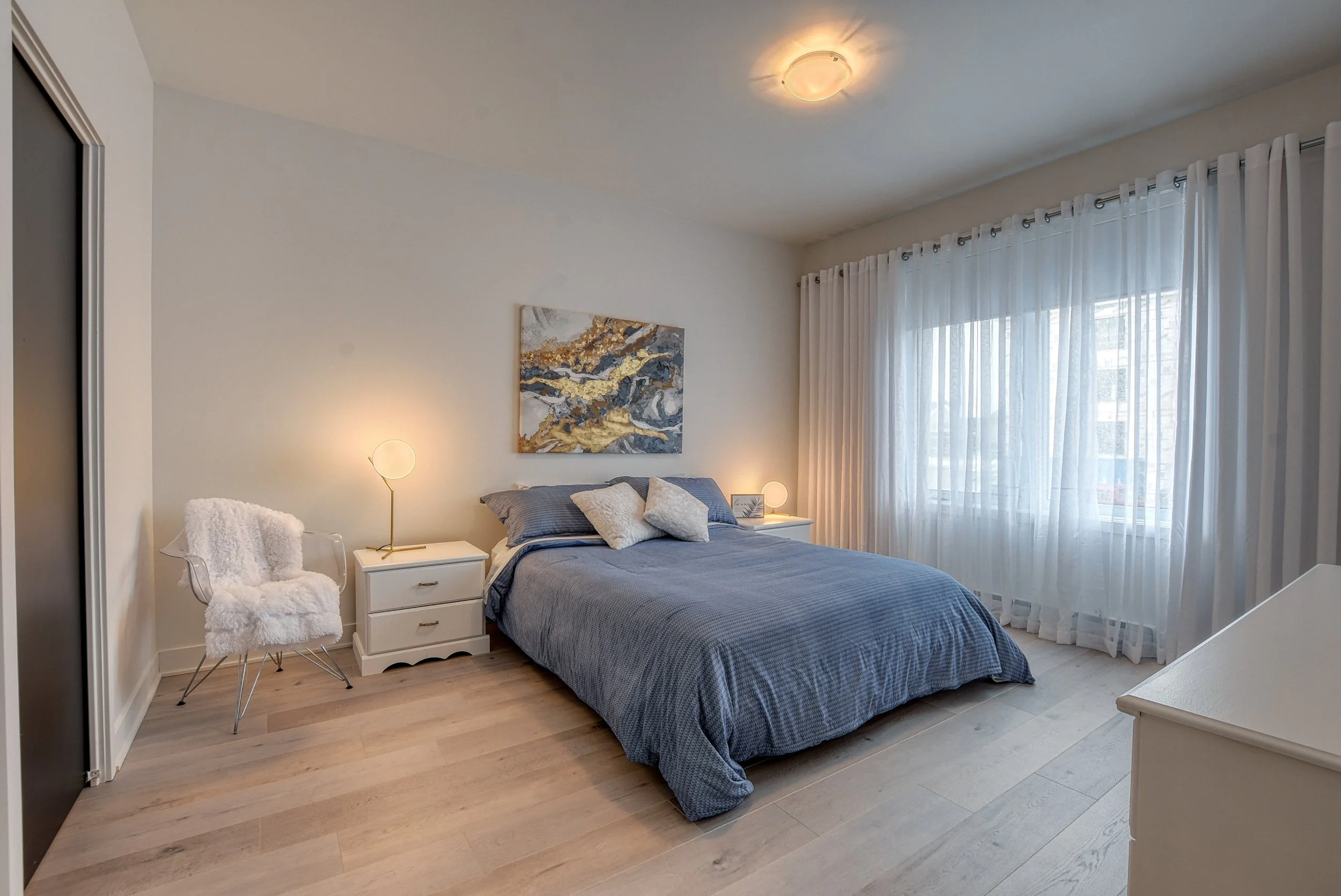 Chambre minimaliste avec un lit en bois clair, draps gris, deux tables de chevet avec lampes, une chaise blanche avec un coussin en peluche, tableau abstrait au mur et grande fenêtre avec rideaux blancs.