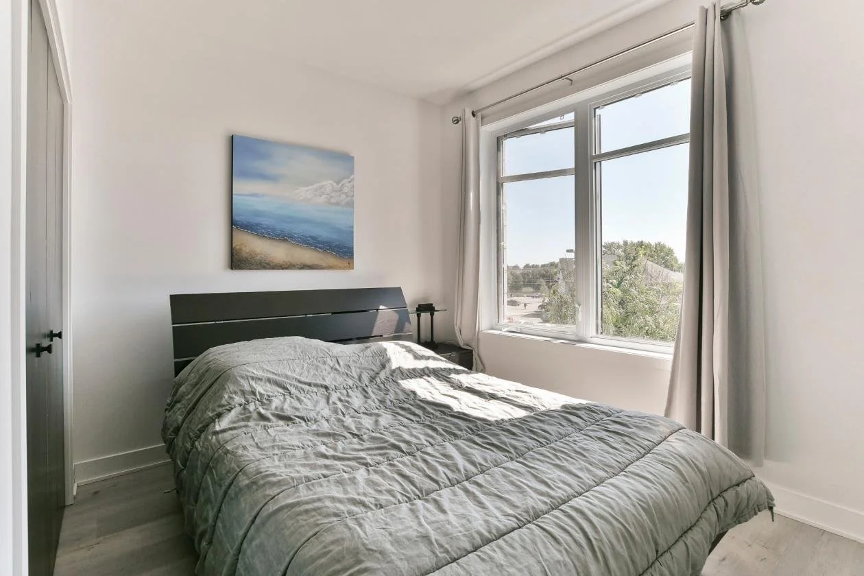 Chambre avec lit à côté d'une grande fenêtre laissant entrer la lumière naturelle, tableau de paysage marin au mur, rideaux gris clair, décoration minimaliste.