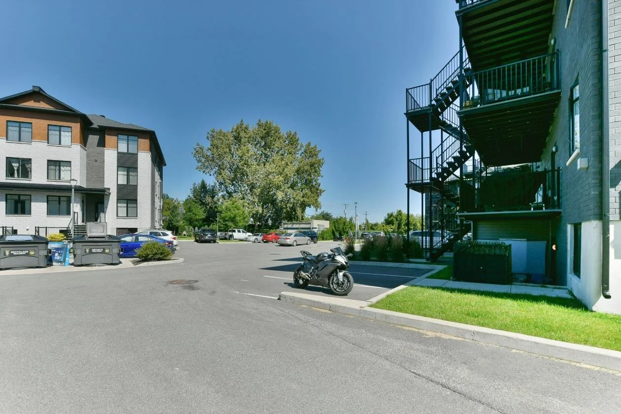 Un parking résidentiel avec plusieurs voitures, un scooter, un bâtiment avec des escaliers en spirale en métal noir, arbres et un bâtiment d'appartements à proximité, ciel clair.