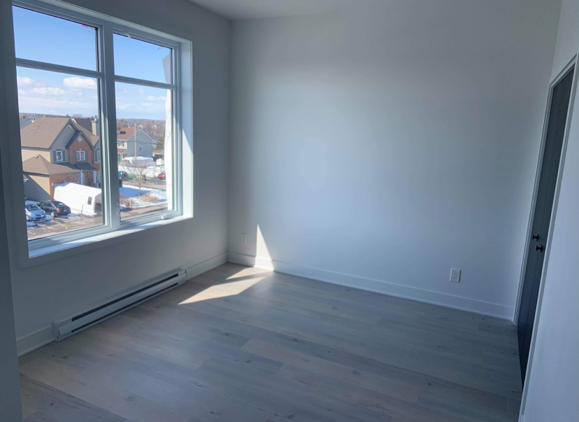 Une pièce vide avec une grande fenêtre laissant entrer de la lumière naturelle, vue sur des maisons et un ciel bleu.