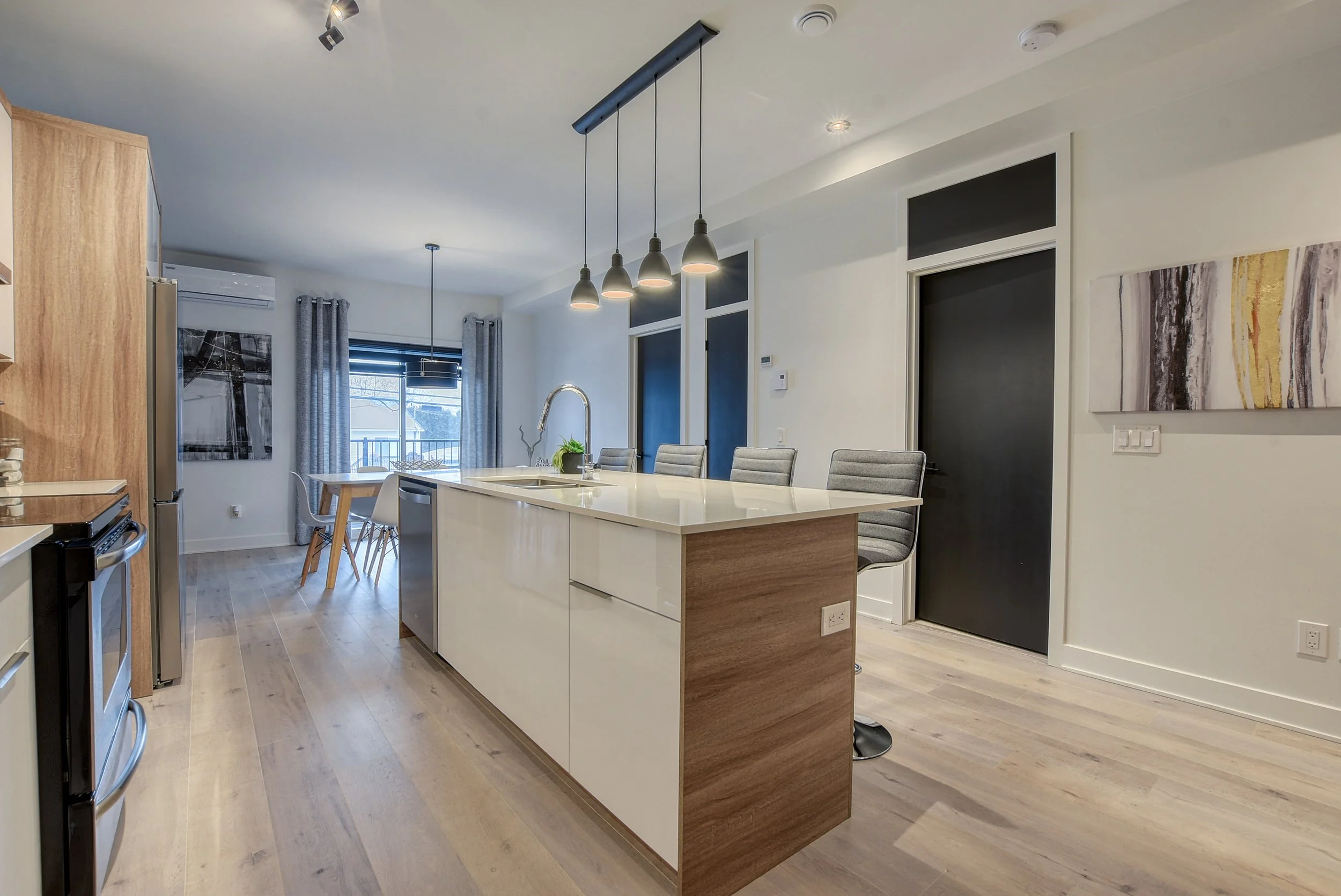 Cuisine moderne avec îlot central en bois et blanc, éclairage suspendu, fenêtres avec rideaux gris, tableaux abstraits sur les murs, sol en bois clair, espace lumineux.