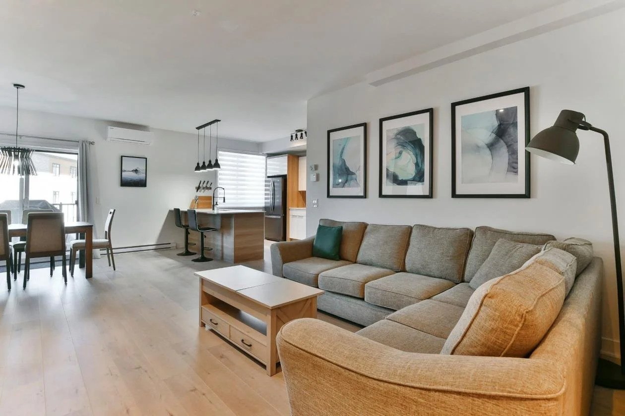 Salon moderne avec canapé beige, tableau abstrait au mur, lampe de lecture, et espace de repas avec meubles en bois, cuisine ouverte en arrière-plan, luminosité naturelle.