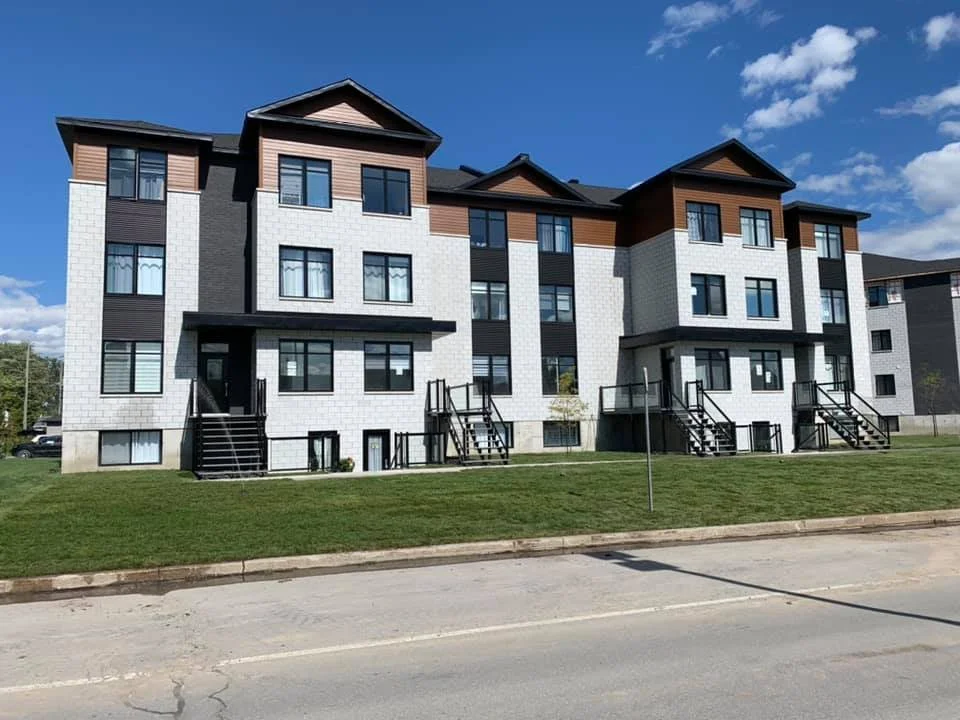 Immeuble résidentiel moderne avec plusieurs appartements, escaliers extérieurs, balcon, et un espace vert devant, sous un ciel bleu avec quelques nuages.