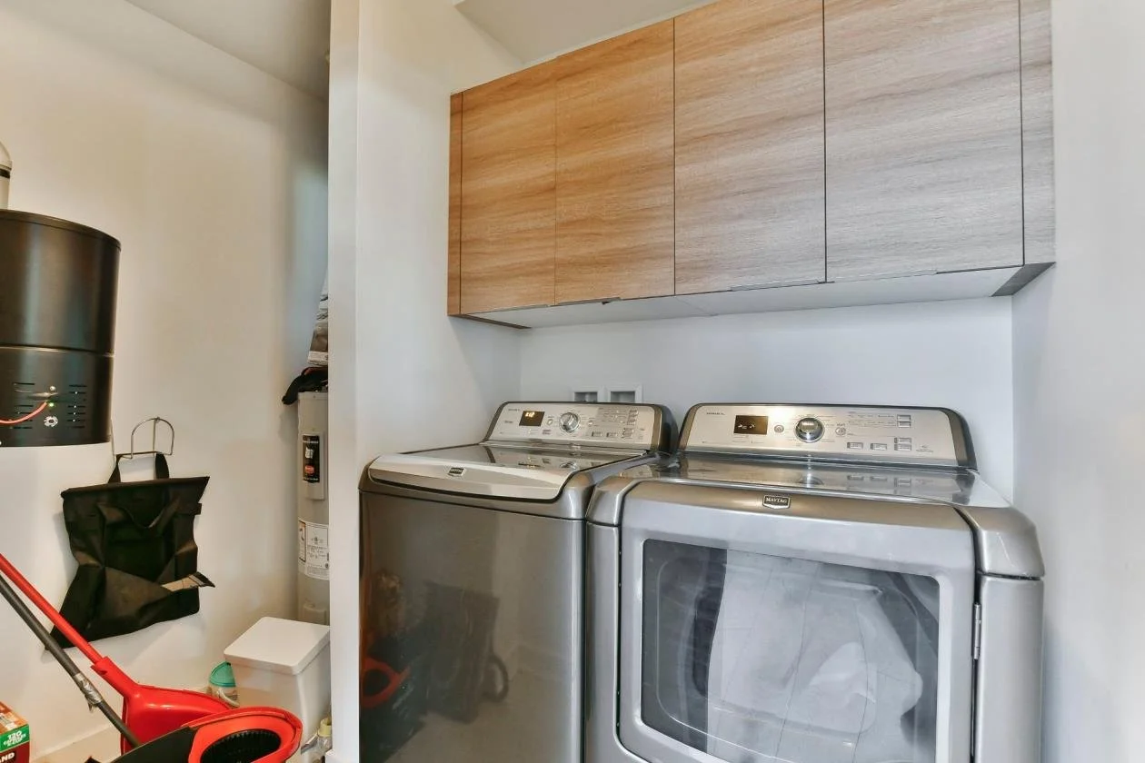 Laverie avec une machine à laver et un sèche-linge en acier inoxydable, des armoires en bois au mur, un balai rouge, un seau noir, un thermos noir, et un petit récipient blanc.