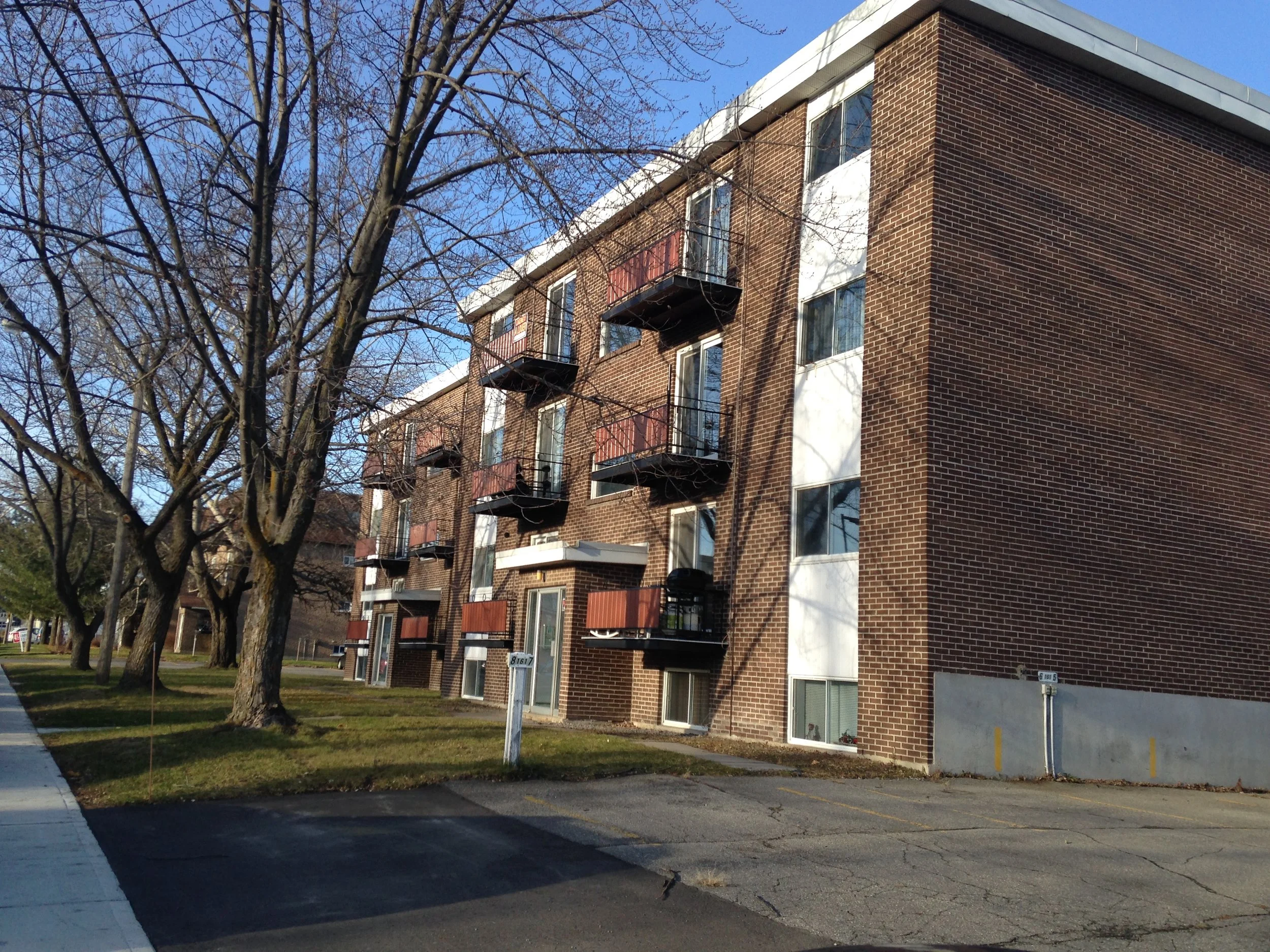 Bâtiment résidentiel en briques avec plusieurs balcons noirs et fenêtres, arbres sans feuilles en premier plan, ciel bleu clair et ombres projetées par le soleil.