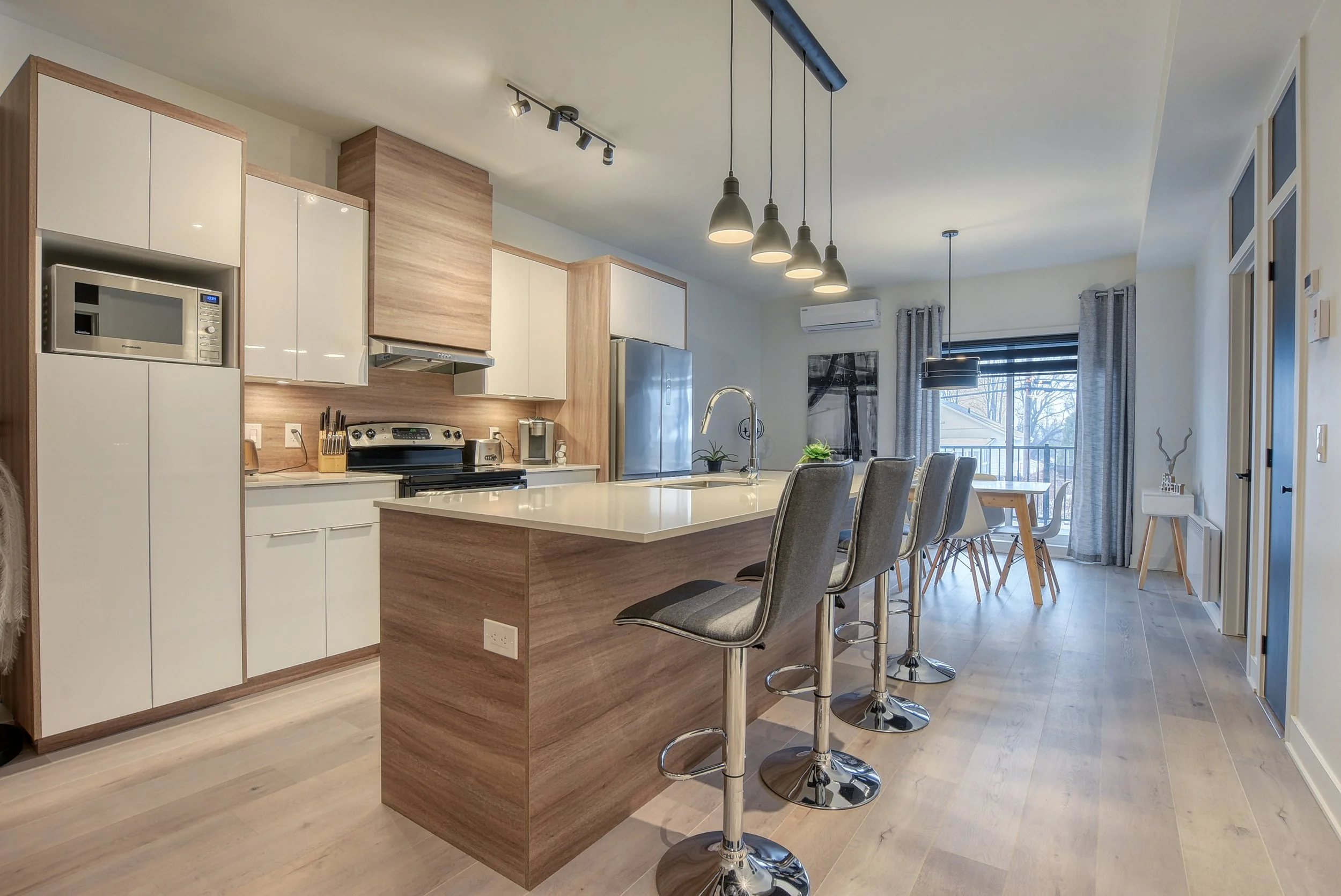 Cuisine moderne avec îlot central en bois et blanc, chaises de bar, table à manger près de la fenêtre, luminaires suspendus, fenêtres avec rideaux, parquet clair.
