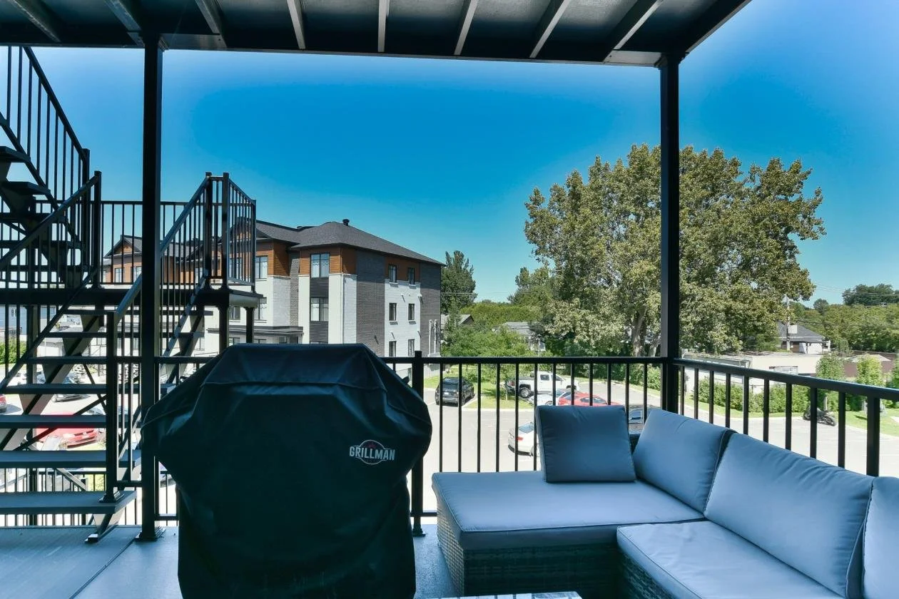 Balcon avec un canapé en cuir gris, un barbecue couvert, et une vue d'un parking avec plusieurs voitures, des arbres et des bâtiments résidentiels, sous un ciel clair.