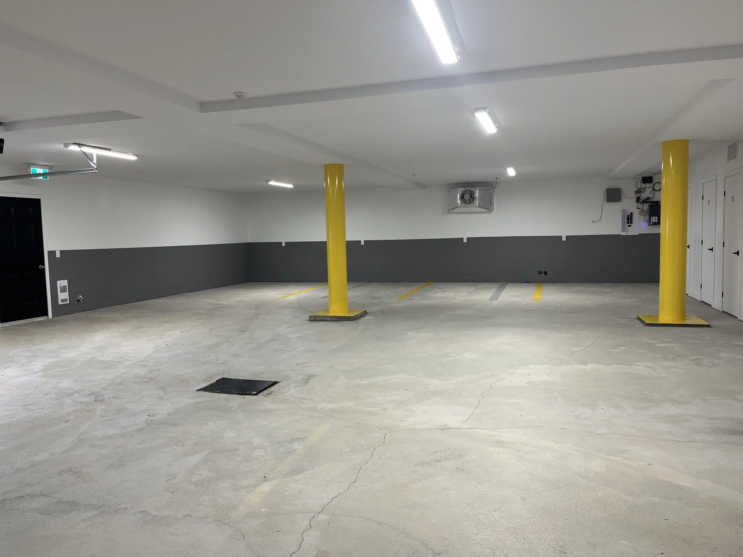 Parking souterrain vide avec deux colonnes jaunes, murs gris et blancs, éclairage fluorescent, et quelques équipements électriques sur la droite.