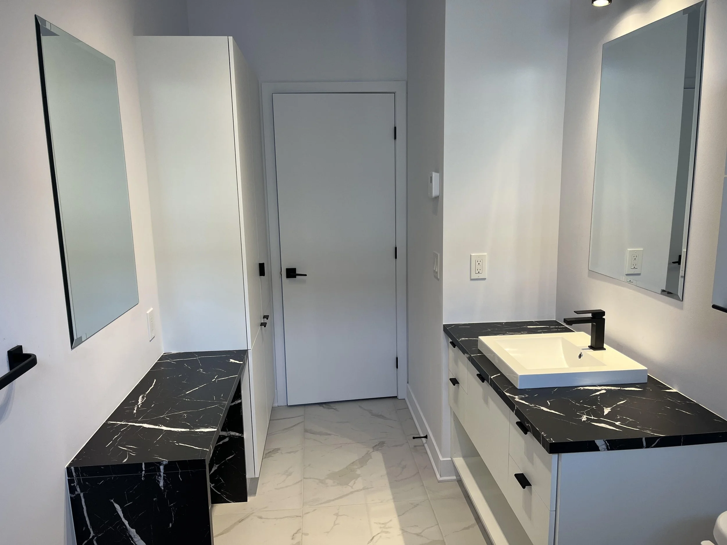 Salle de bain moderne avec deux lavabos, miroirs, armoires blanches, comptoirs en marbre noir, carrelage blanc et un sèche-serviettes noir.