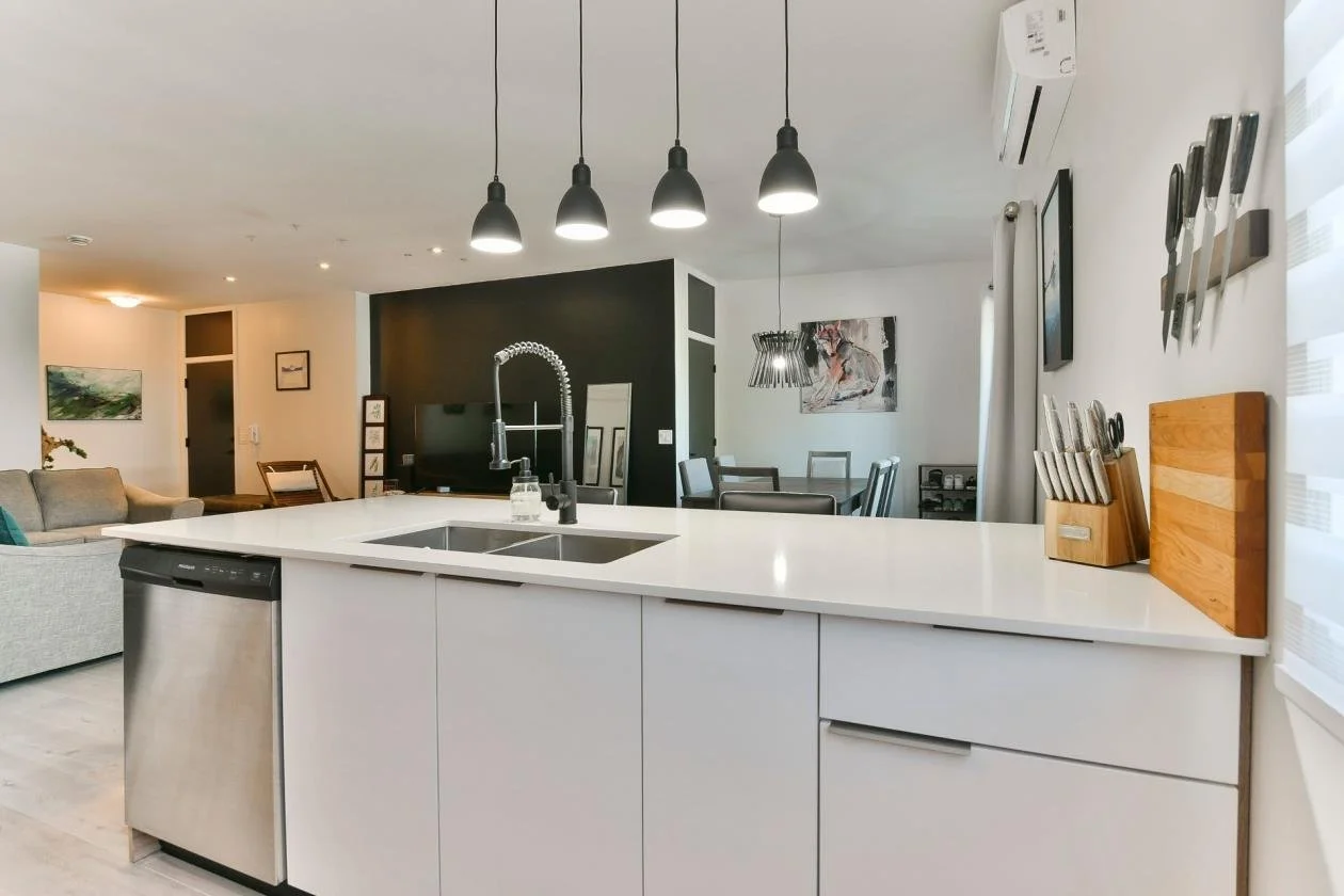 Cuisine moderne avec un plan de travail blanc, un évier à évier creux, un robinet spiralé, un lave-vaisselle intégré, un tableau de couteaux en bois, des lampes suspendues, et un salon avec mobilier en toile beige, tableau et éléments décoratifs vari