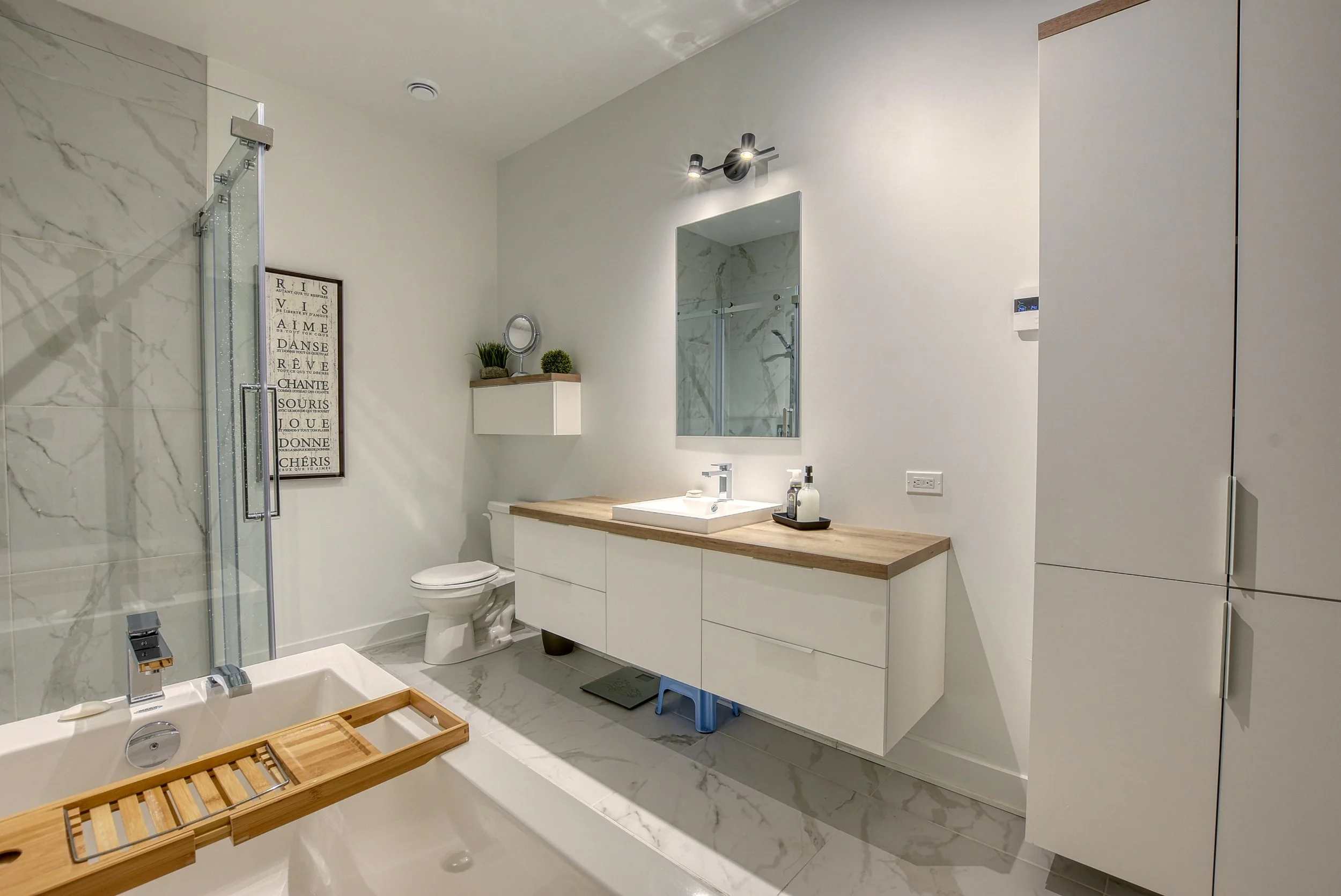 Salle de bain moderne avec douche en verre, baignoire, miroir, lavabo sur meuble blanc en bois, toilette, armoires de rangement, décoration minimaliste, plantes et affiches