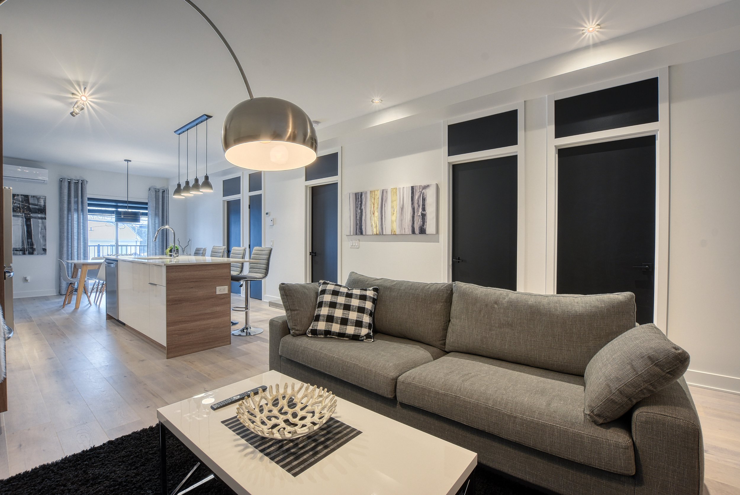 Intérieur d'un salon moderne avec un canapé gris, une table basse blanche, et une zone de cuisine avec îlot et chaises.